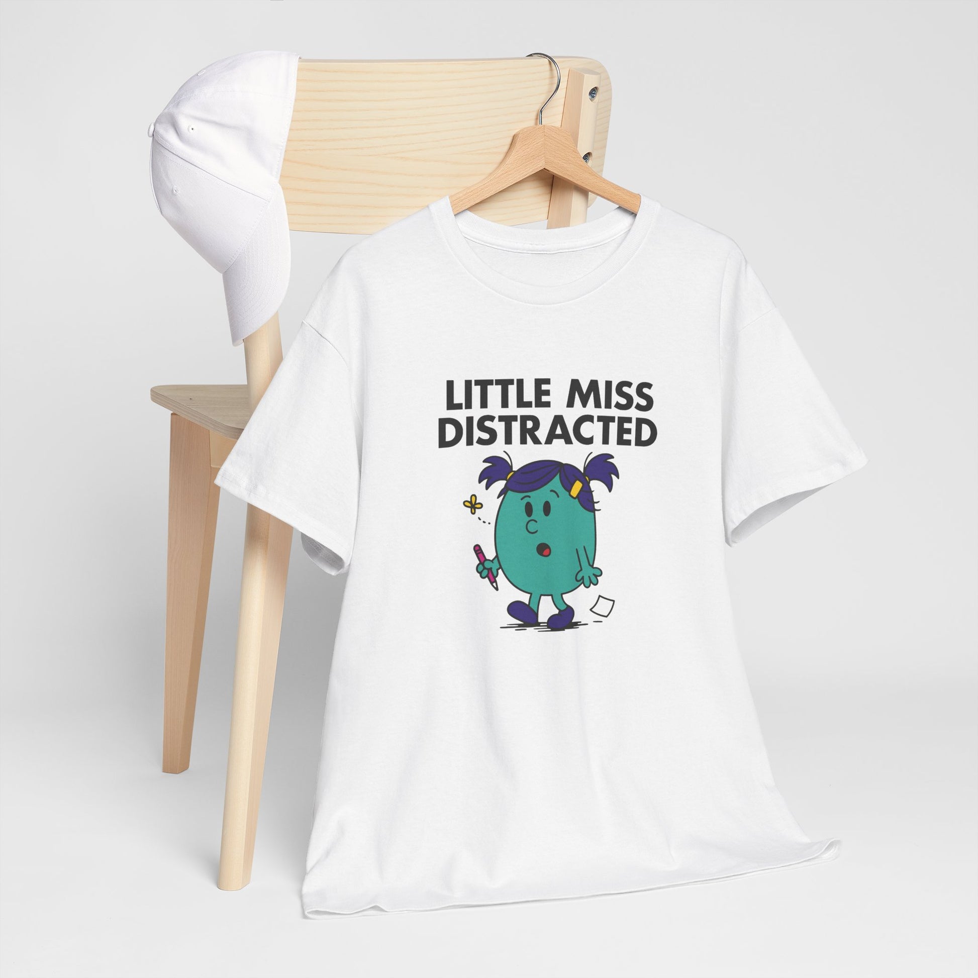 Little Miss Distracted T-shirt draped over a wooden chair in a casual setting. Part of the Little Miss Meme Character Collection. A Vivid Divergence Sensory Friendly Unisex Tee.

