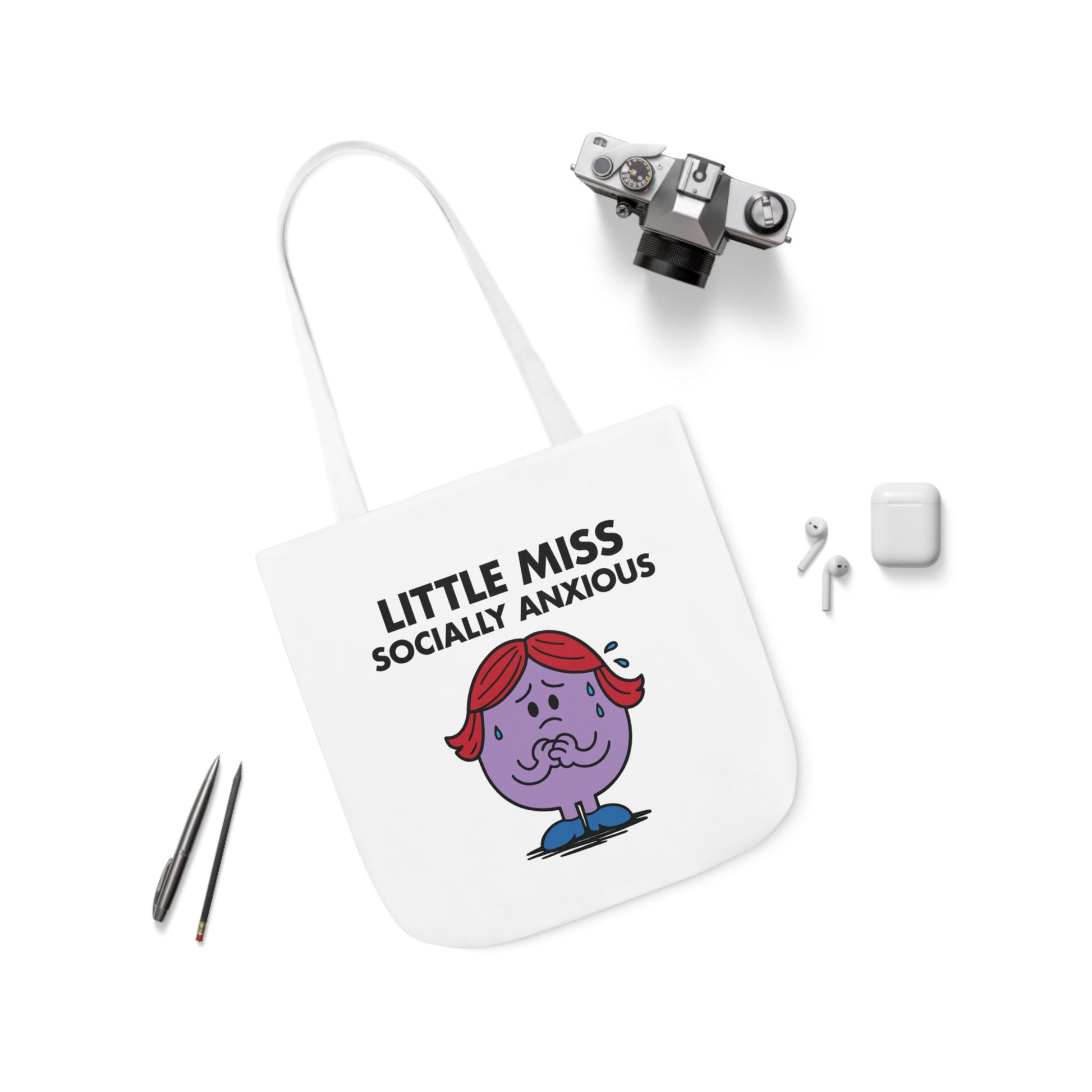 Flat lay of tote bag with Little Miss Socially Anxious surrounded by earbuds and pens. She’s recharging after replying “you too” to the waiter. Part of the Vivid Divergence Little Miss Meme Collection!
