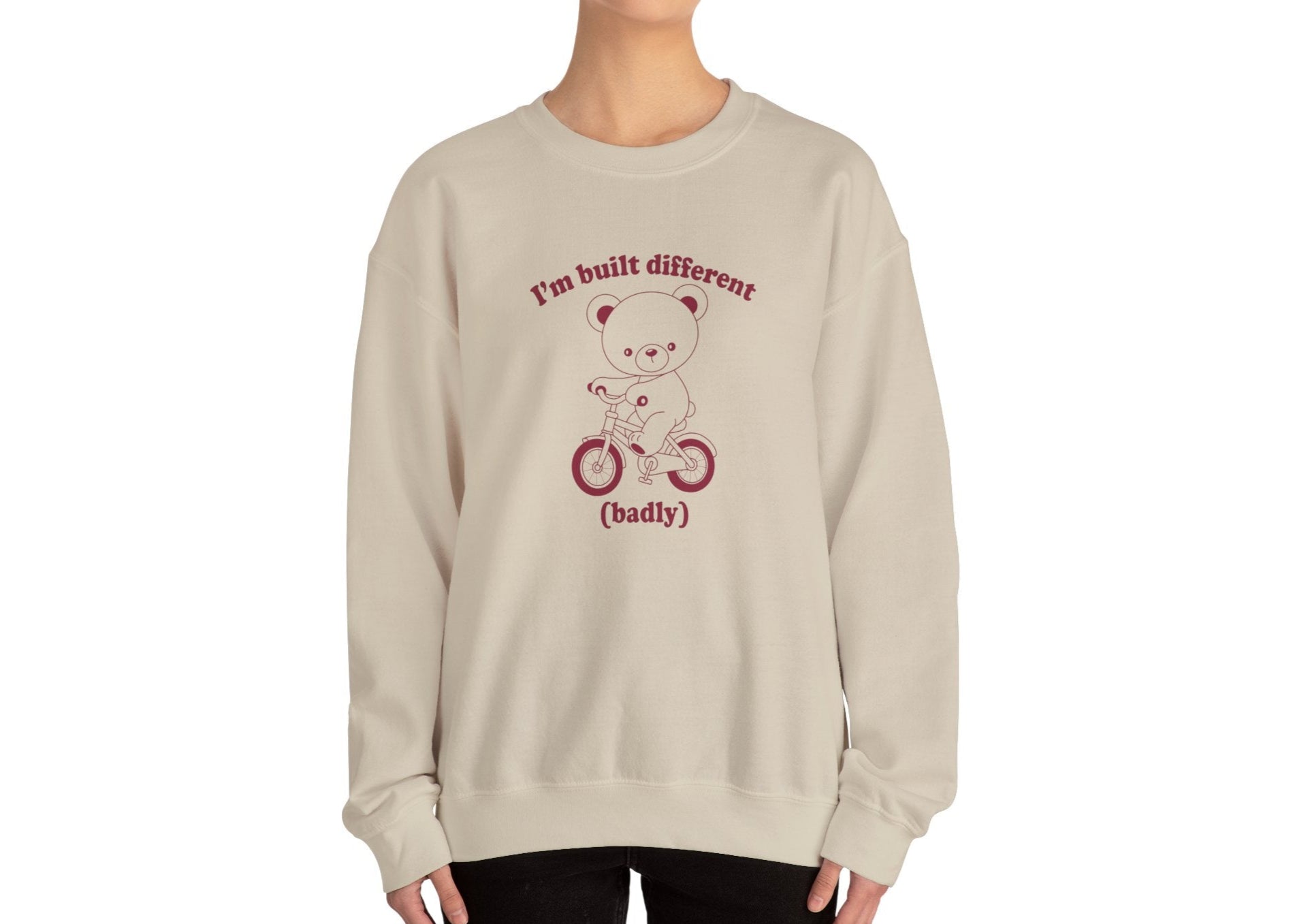 Model wearing a beige sweatshirt with a graphic of a bear riding a bike and the text "I'm Built Different (Badly)," standing with hands at sides. Part of the Vivid Divergence Sensory Friendly Unisex Sweater Jumper Range.