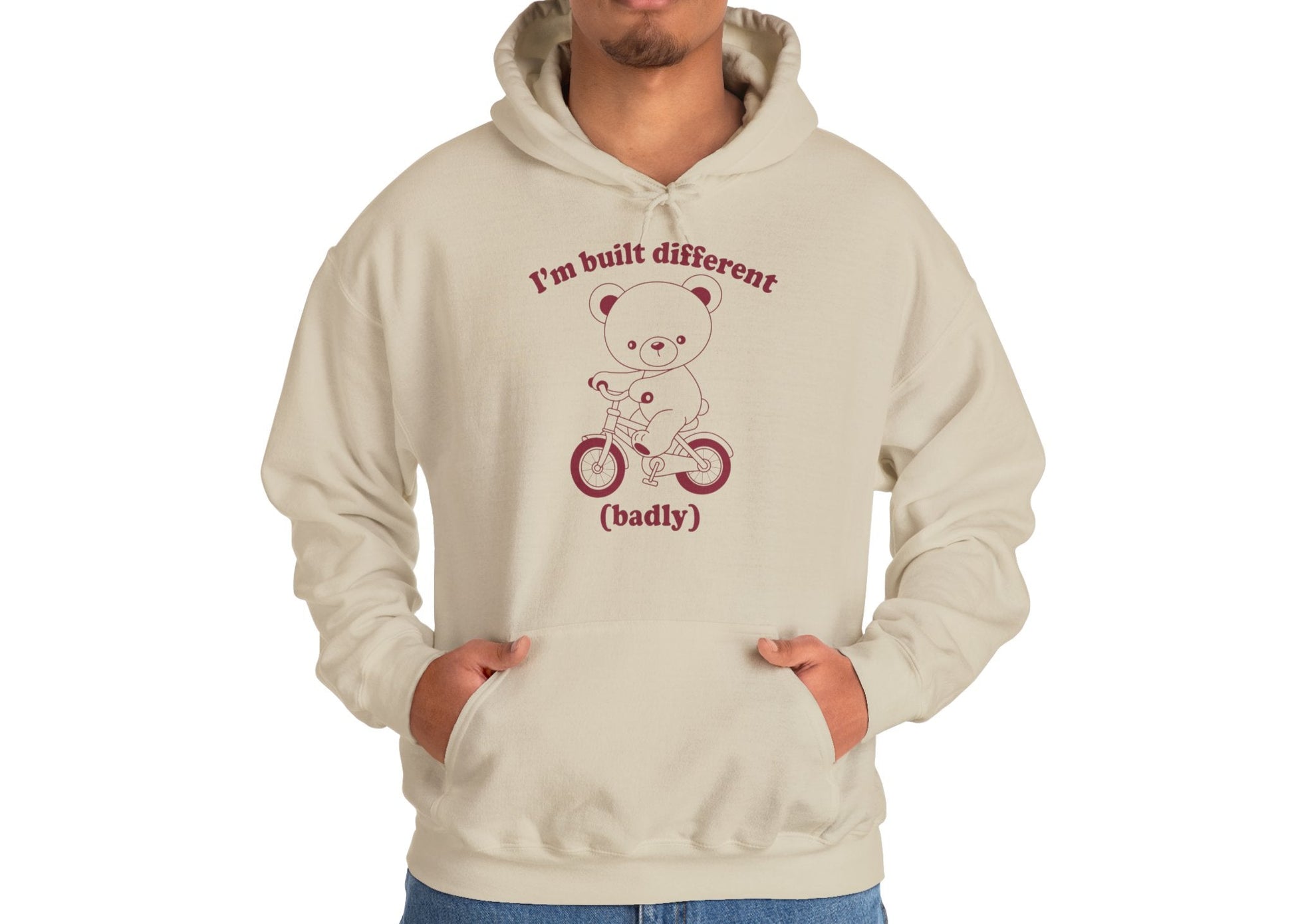 Model wearing a beige hoodie with a cartoon bear riding a small bicycle and the text "I'm Built Different (Badly)," standing with hands in pockets. Part of the Vivid Divergence Sensory Friendly Unisex Hoodie Range.