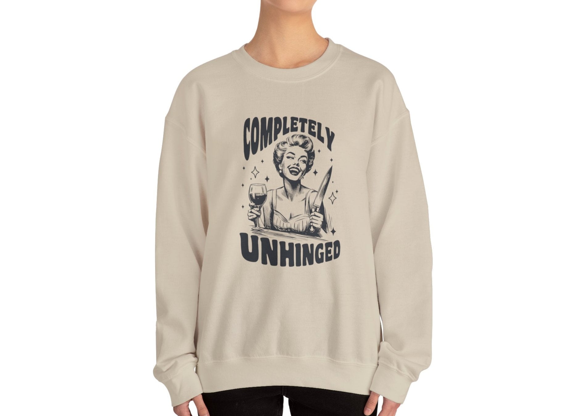 Model wearing a beige sweatshirt with a graphic of a woman holding a knife and a glass of wine and the text "Completely Unhinged," standing with hands at sides. Part of the Vivid Divergence Sensory Friendly Unisex Sweater Jumper Range.