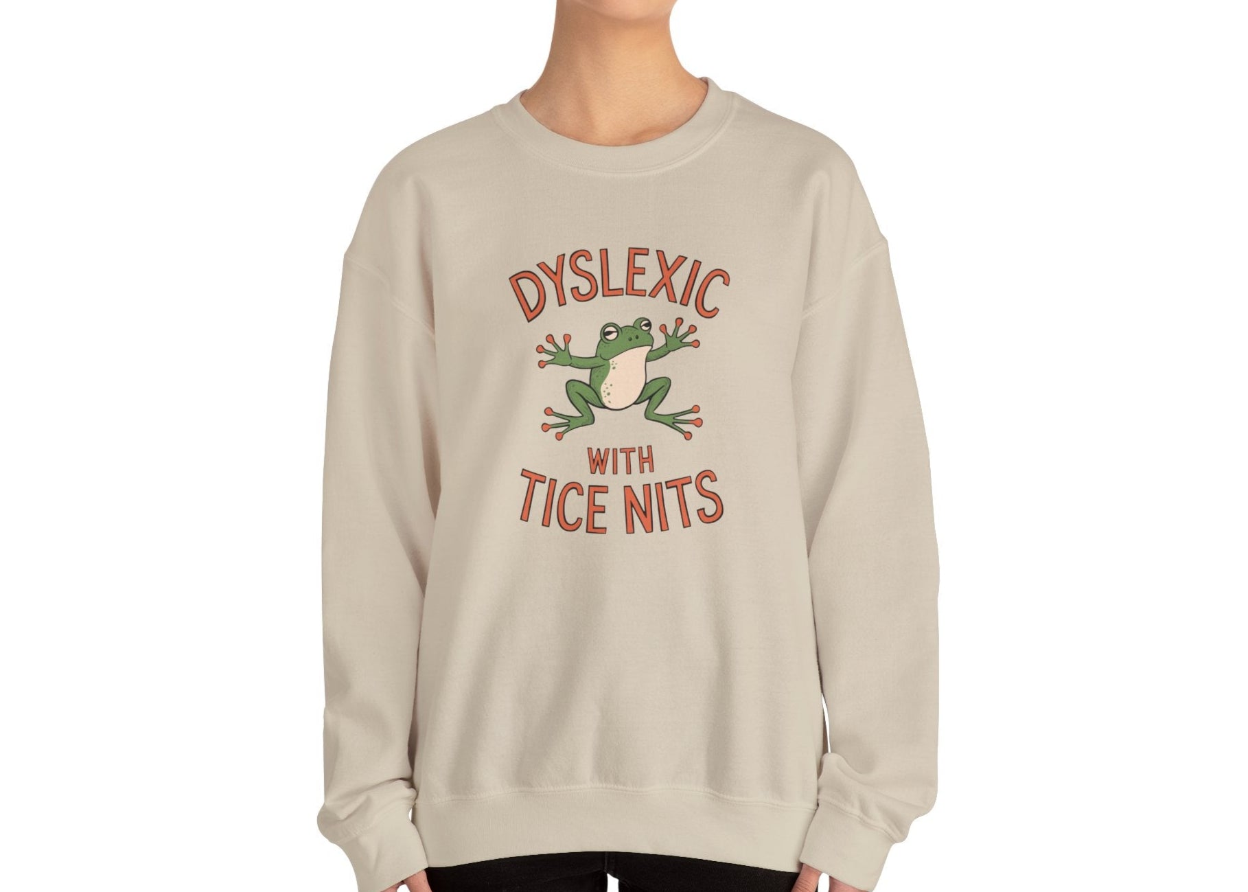 Model wearing a beige sweatshirt with a cartoon frog and the text "Dyslexic with Tice Nits," standing with hands at sides. Part of the Vivid Divergence Sensory Friendly Unisex Sweater Jumper Range.