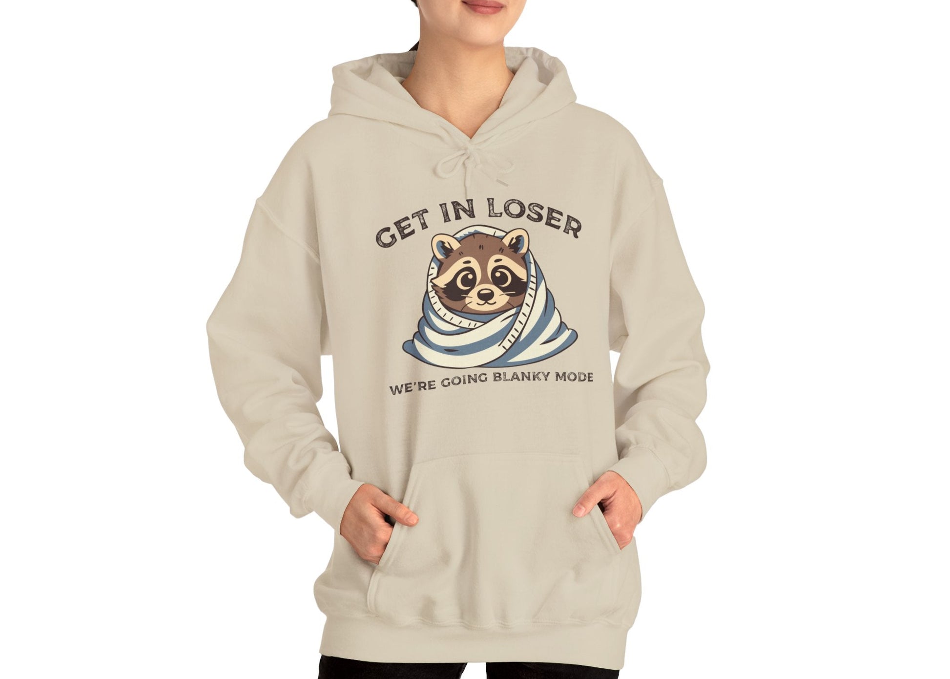 Model wearing a beige hoodie with a raccoon wrapped in a blanket and the text "Get in Loser, We're Going Blanky Mode," standing with hands in pockets. Part of the Vivid Divergence Sensory Friendly Unisex Hoodie Range.