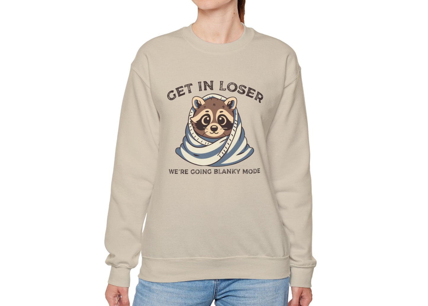 Model wearing a beige sweatshirt with a raccoon wrapped in a blanket and the text "Get in Loser, We're Going Blanky Mode," standing with hands at sides. Part of the Vivid Divergence Sensory Friendly Unisex Sweater Jumper Range.