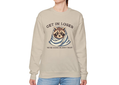 Model wearing a beige sweatshirt with a raccoon wrapped in a blanket and the text "Get in Loser, We're Going Blanky Mode," standing with hands at sides. Part of the Vivid Divergence Sensory Friendly Unisex Sweater Jumper Range.
