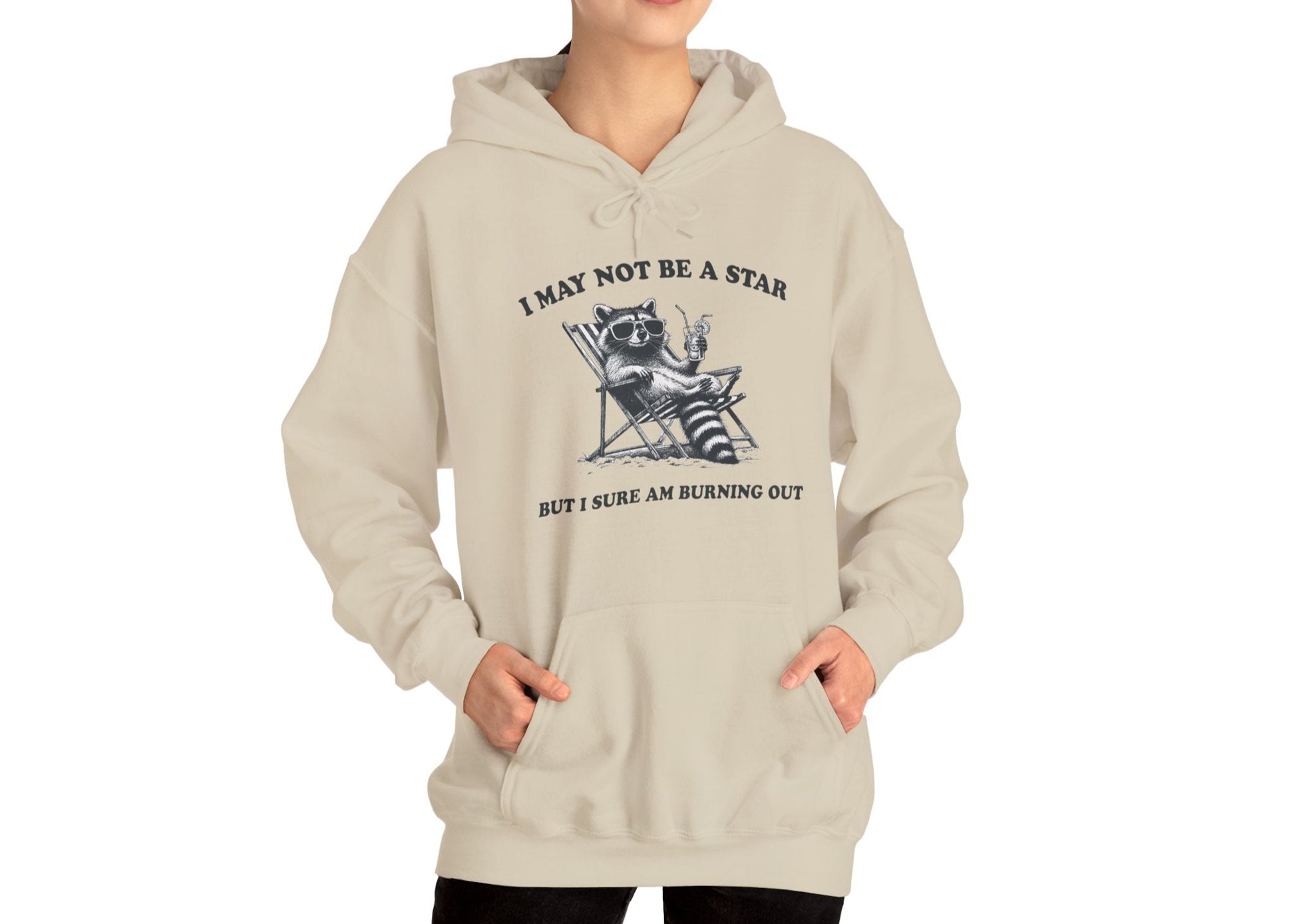 Model wearing a beige hoodie with a raccoon sitting in a trash can and the text "I May Not Be A Star, But I Sure Am Burning Out," standing with hands in pockets. Part of the Vivid Divergence Sensory Friendly Unisex Hoodie Range.