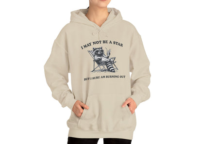 Model wearing a beige hoodie with a raccoon sitting in a trash can and the text "I May Not Be A Star, But I Sure Am Burning Out," standing with hands in pockets. Part of the Vivid Divergence Sensory Friendly Unisex Hoodie Range.