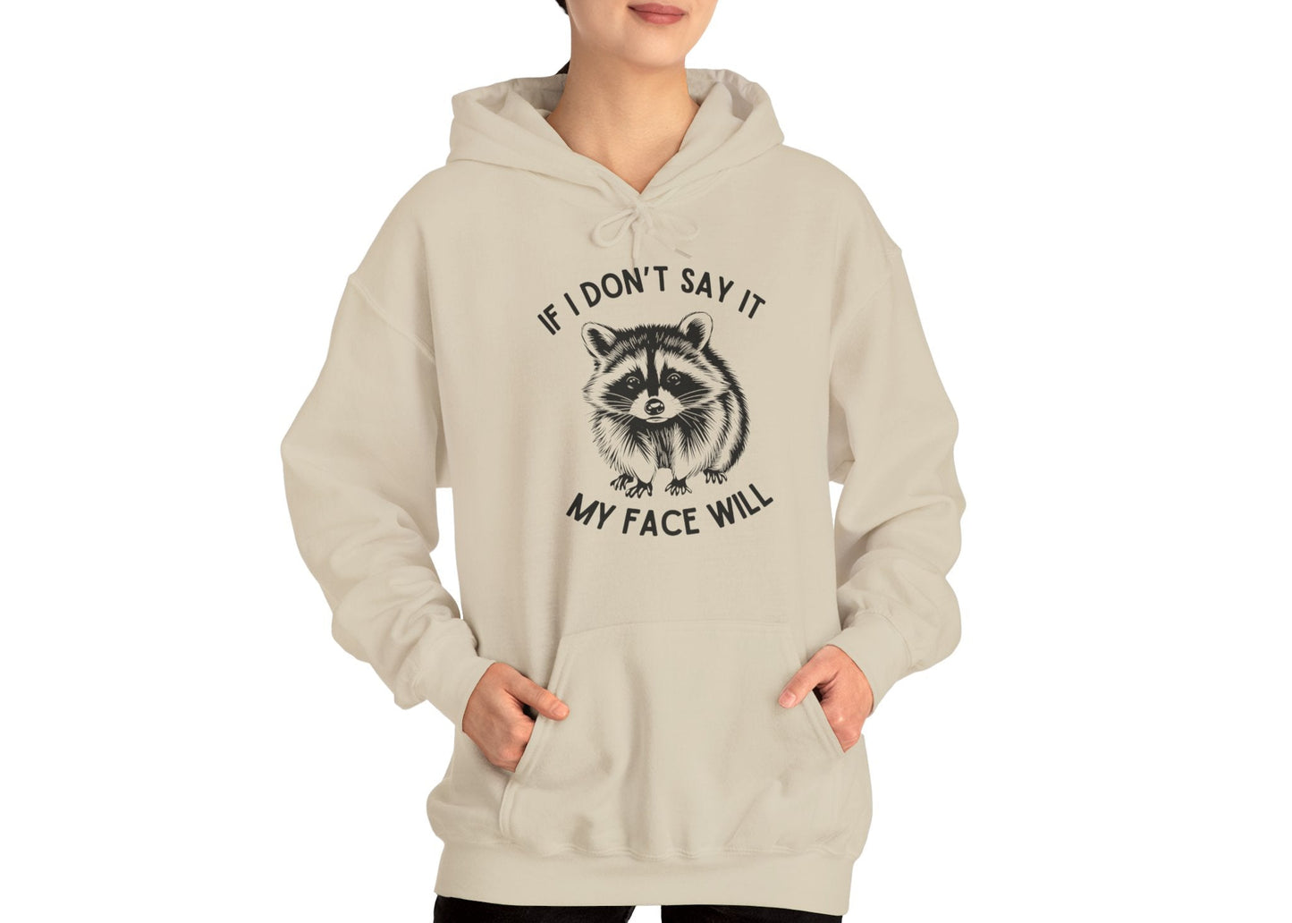 Model wearing a beige hoodie with a raccoon graphic and the text "If I Don't Say It, My Face Will," standing with hands in pockets. Part of the Vivid Divergence Sensory Friendly Unisex Hoodie Range.