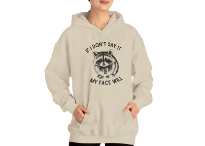 Model wearing a beige hoodie with a raccoon graphic and the text "If I Don't Say It, My Face Will," standing with hands in pockets. Part of the Vivid Divergence Sensory Friendly Unisex Hoodie Range.