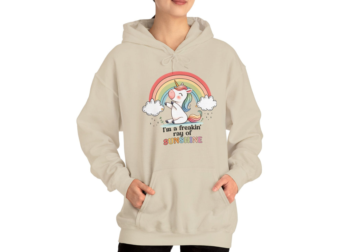 Model wearing a beige hoodie with a grumpy-looking unicorn under a rainbow and the text "I'm a Freakin' Ray of Sunshine," standing with hands in pockets. Part of the Vivid Divergence Sensory Friendly Unisex Hoodie Range.