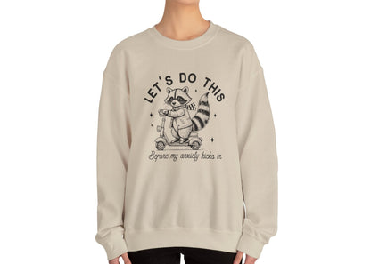 Model wearing a beige sweatshirt with a cartoon raccoon riding a scooter and the text "Let's Do This Before My Anxiety Kicks In," standing with hands at sides. Part of the Vivid Divergence Sensory Friendly Unisex Sweater Jumper Range.