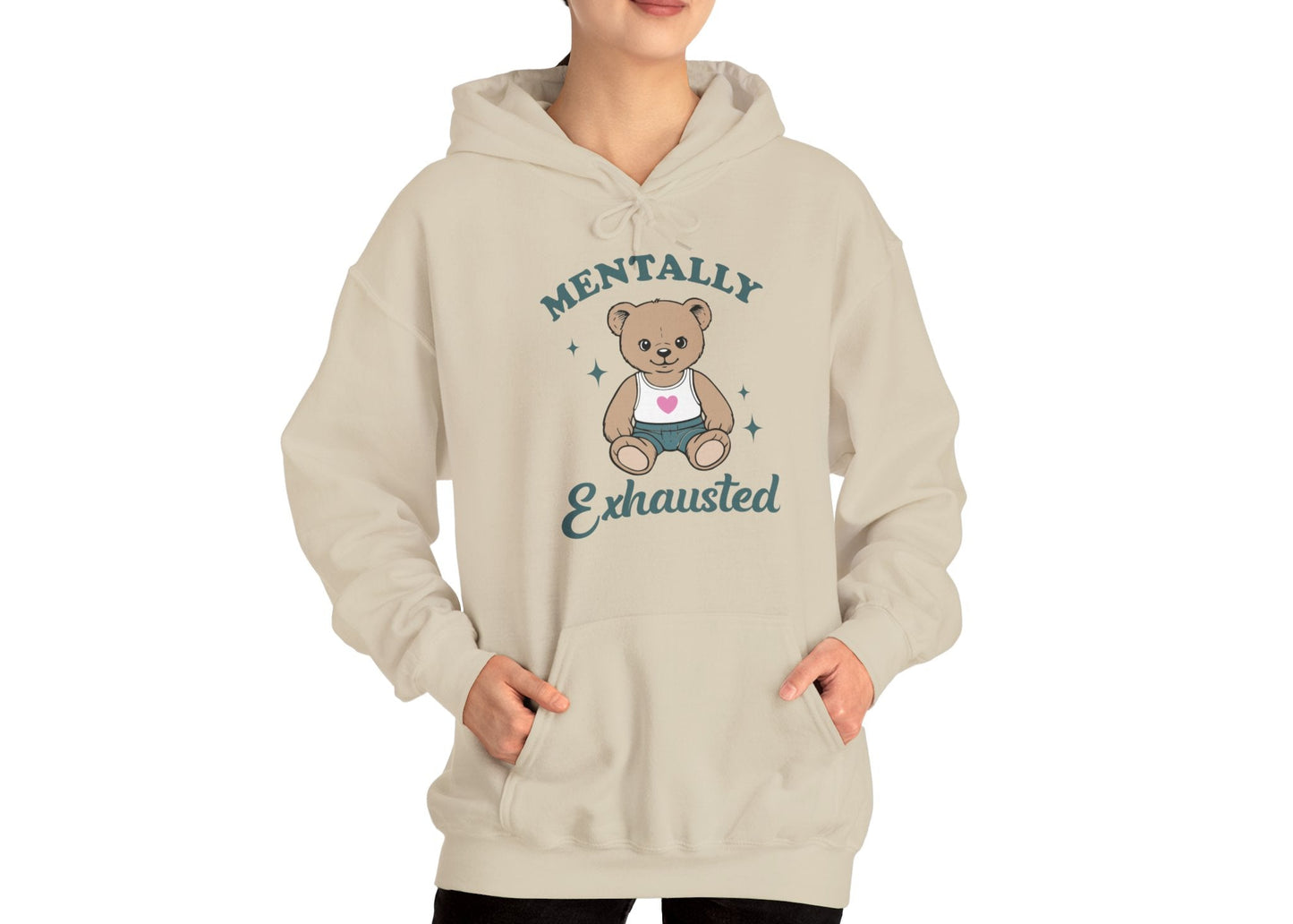 Model wearing a beige hoodie with a cute teddy bear wearing an apron and the text "Mentally Exhausted," standing with hands in pockets. Part of the Vivid Divergence Sensory Friendly Unisex Hoodie Range.