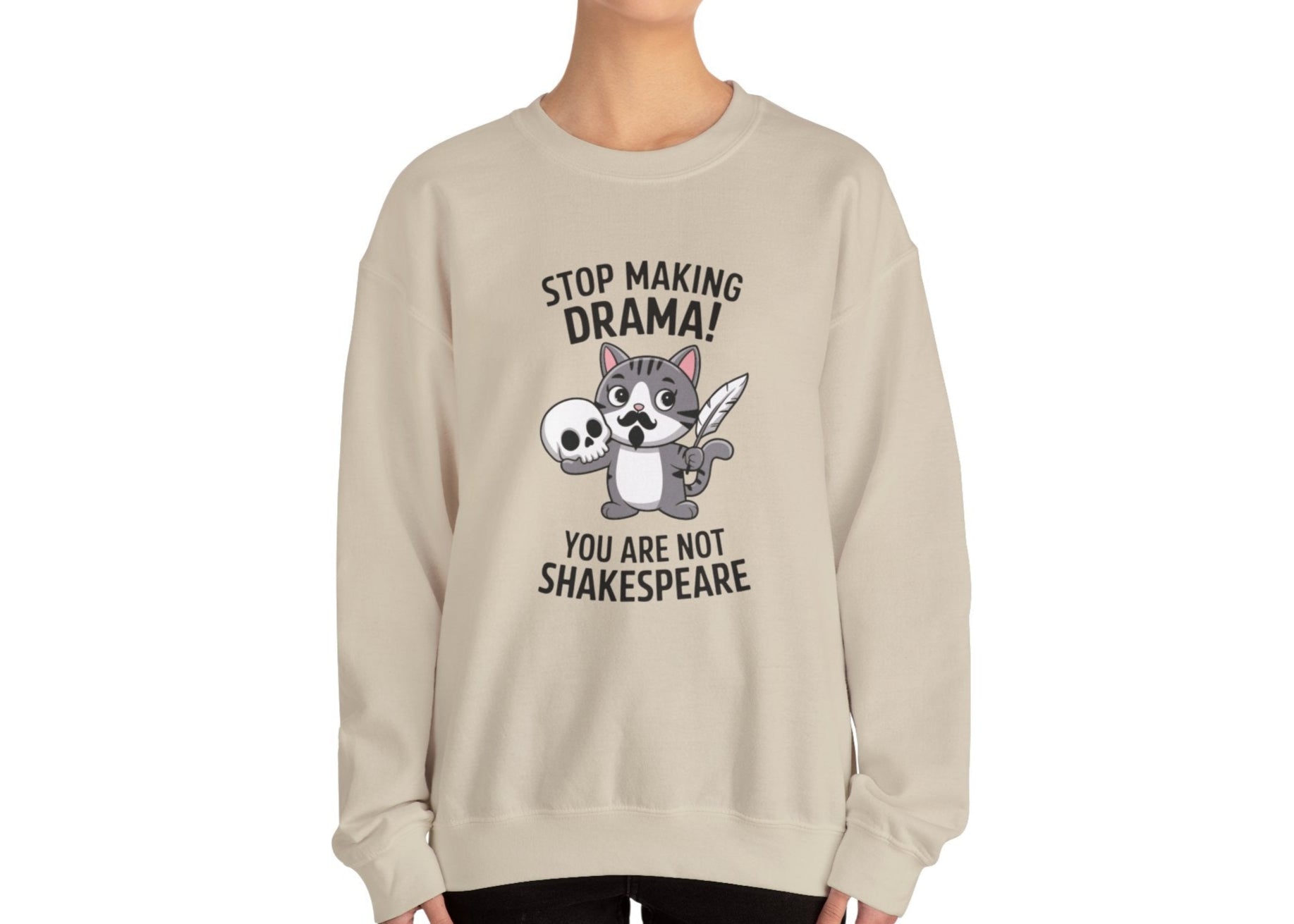 Model wearing a beige sweatshirt with a cartoon cat holding a skull and the text "Stop Making Drama! You Are Not Shakespeare," standing with hands at sides. Part of the Vivid Divergence Sensory Friendly Unisex Sweater Jumper Range.