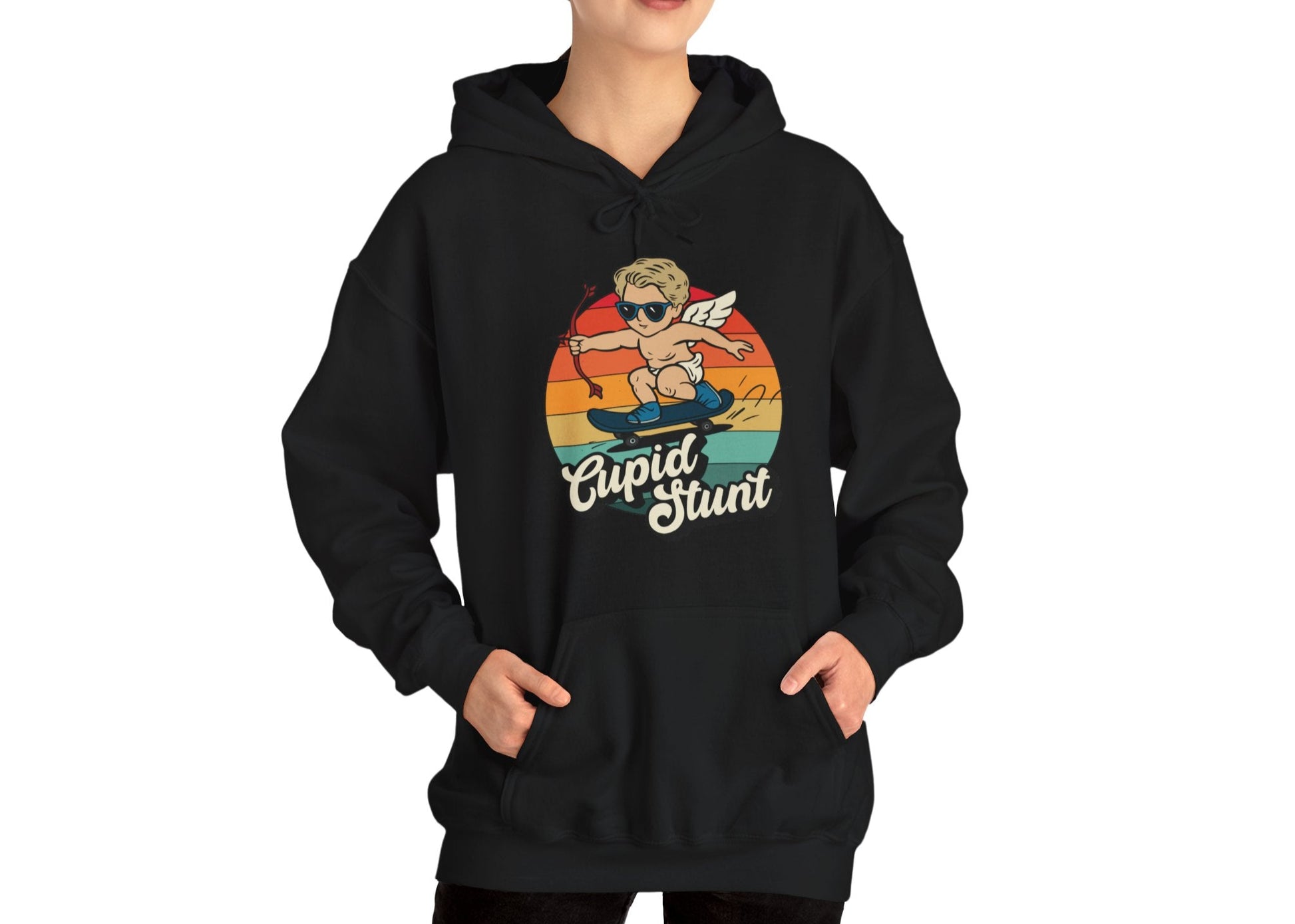 Model wearing a black hoodie with a retro-style sunset background, an illustration of Cupid riding a skateboard, and the text "Cupid Stunt," standing with hands in pockets. Part of the Vivid Divergence Sensory Friendly Unisex Hoodie Range.