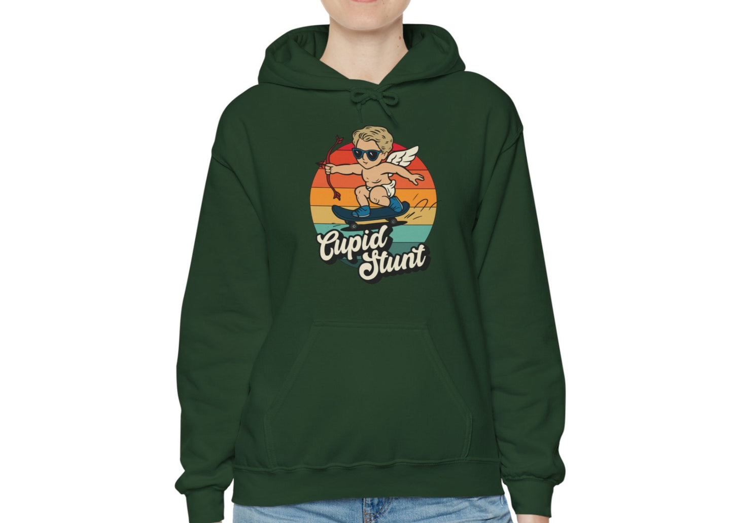 Model wearing a forest green hoodie with a retro-style sunset background, an illustration of Cupid riding a skateboard, and the text "Cupid Stunt," standing with hands at sides. Part of the Vivid Divergence Sensory Friendly Unisex Hoodie Range.