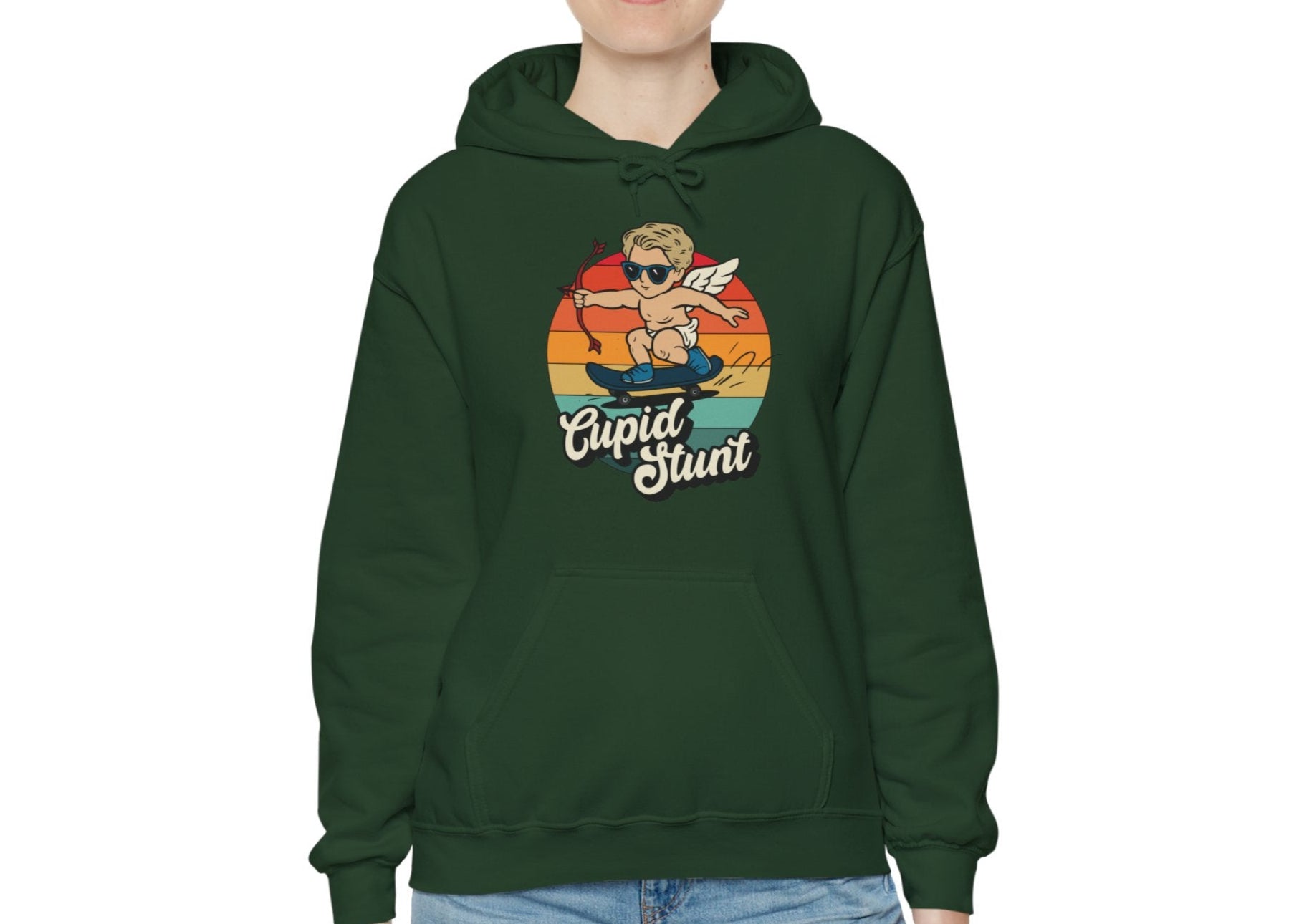 Model wearing a forest green hoodie with a retro-style sunset background, an illustration of Cupid riding a skateboard, and the text "Cupid Stunt," standing with hands at sides. Part of the Vivid Divergence Sensory Friendly Unisex Hoodie Range.