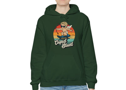 Model wearing a forest green hoodie with a retro-style sunset background, an illustration of Cupid riding a skateboard, and the text "Cupid Stunt," standing with hands at sides. Part of the Vivid Divergence Sensory Friendly Unisex Hoodie Range.