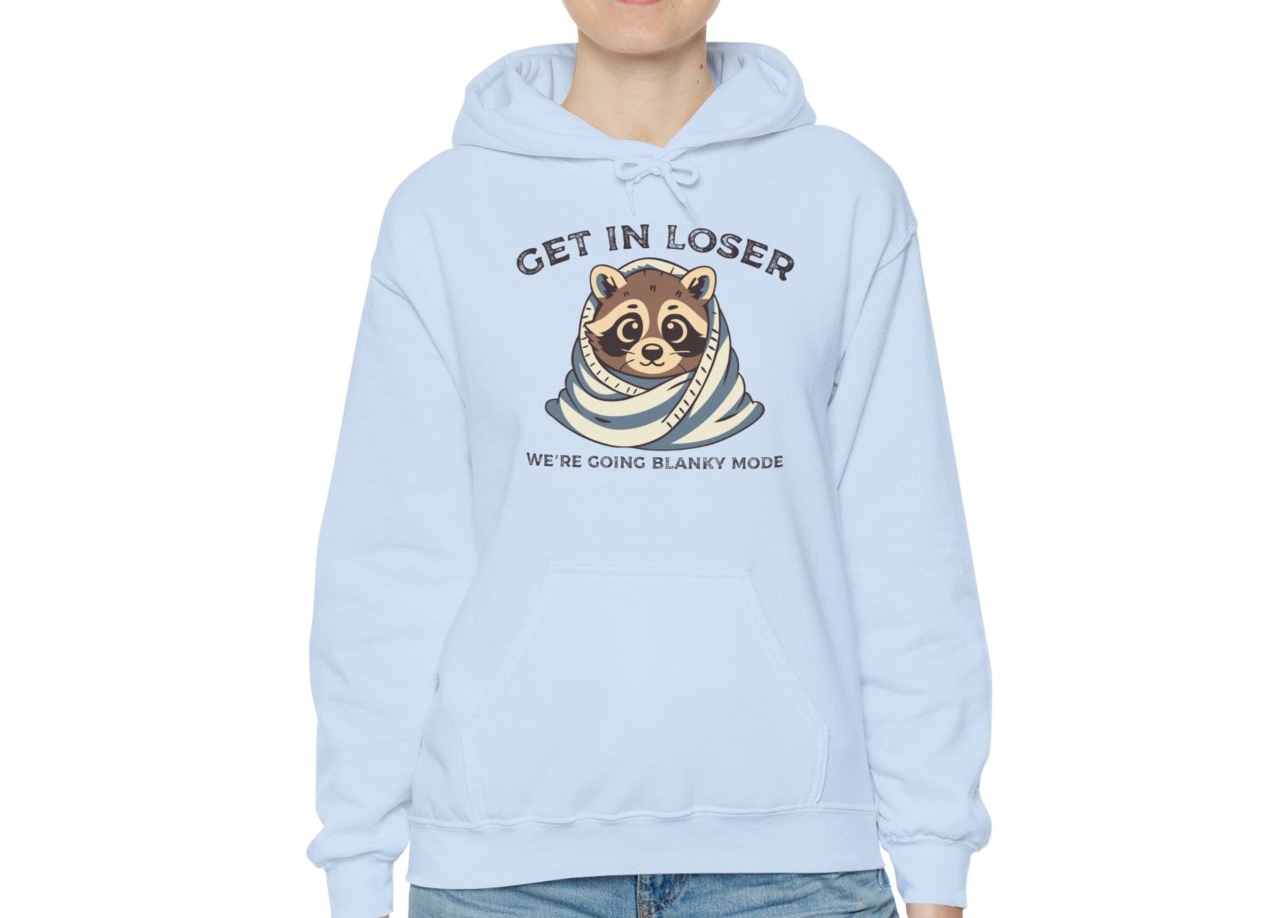 Model wearing a light blue hoodie with a raccoon wrapped in a blanket and the text "Get in Loser, We're Going Blanky Mode," standing with hands at sides. Part of the Vivid Divergence Sensory Friendly Unisex Hoodie Range.