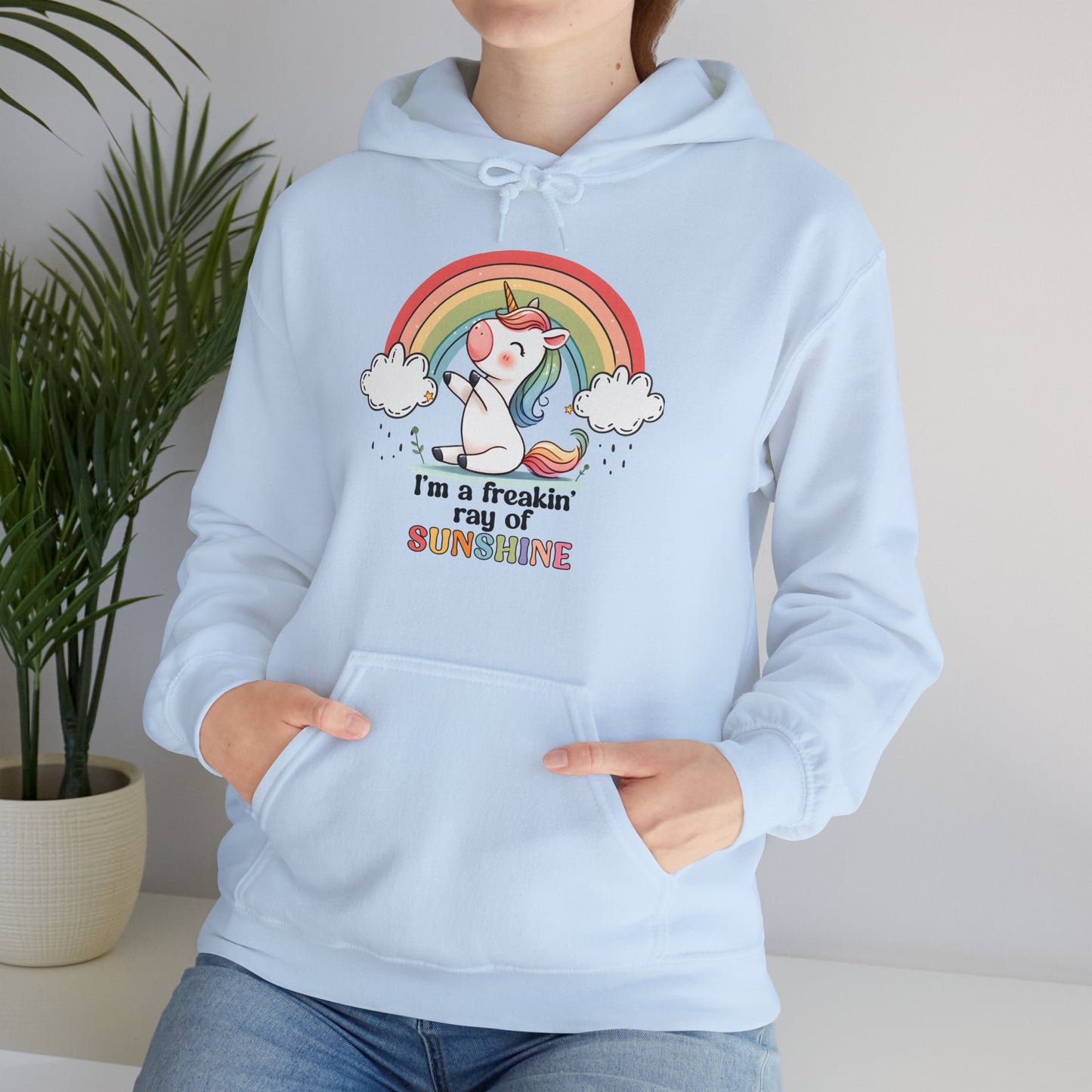 Model wearing a light blue hoodie with a grumpy-looking unicorn under a rainbow and the text "I'm a Freakin' Ray of Sunshine," standing with hands at sides. Part of the Vivid Divergence Sensory Friendly Unisex Hoodie Range.