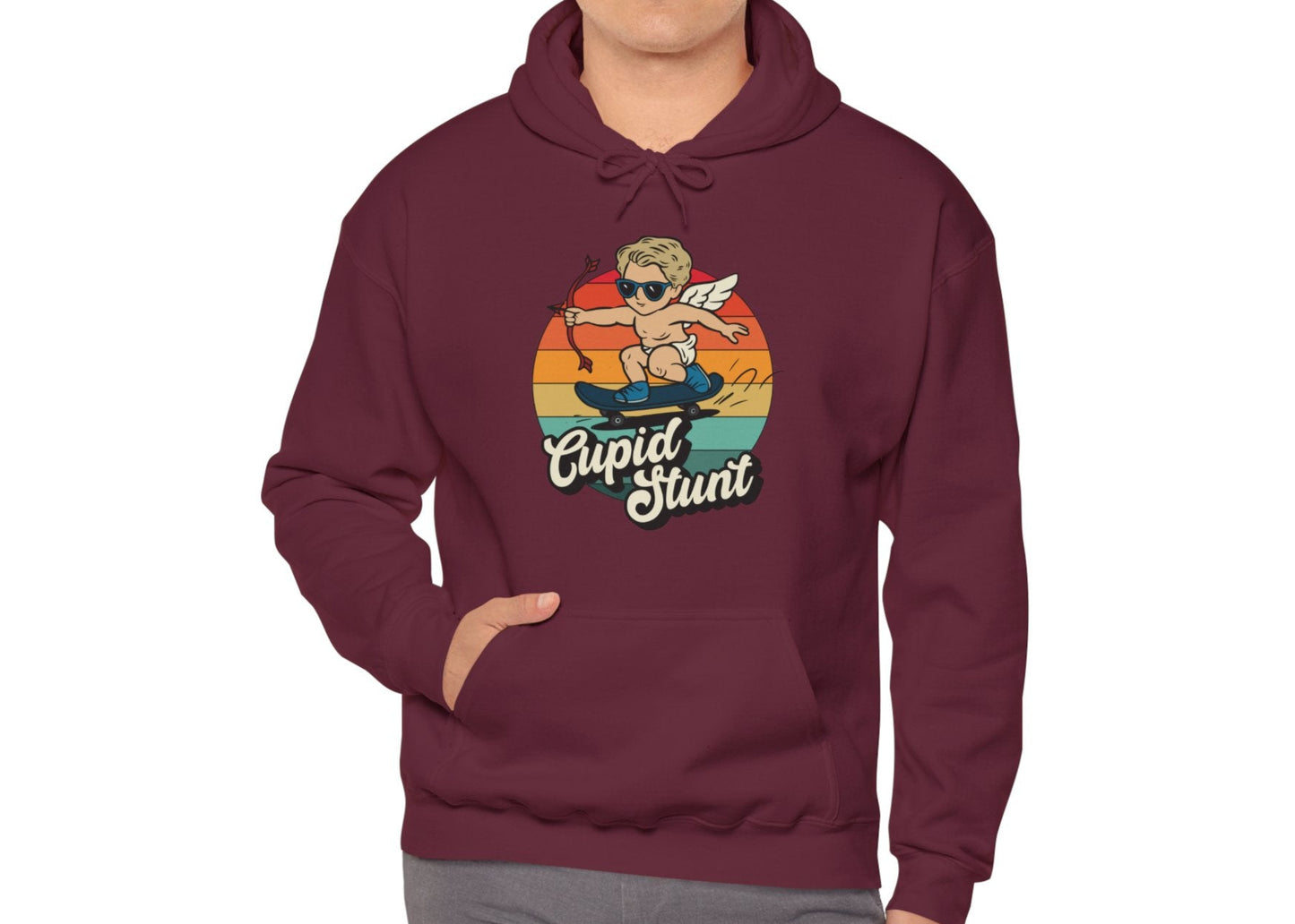 Model wearing a maroon hoodie with a retro-style sunset background, an illustration of Cupid riding a skateboard, and the text "Cupid Stunt," standing with hands in pockets. Part of the Vivid Divergence Sensory Friendly Unisex Hoodie Range.