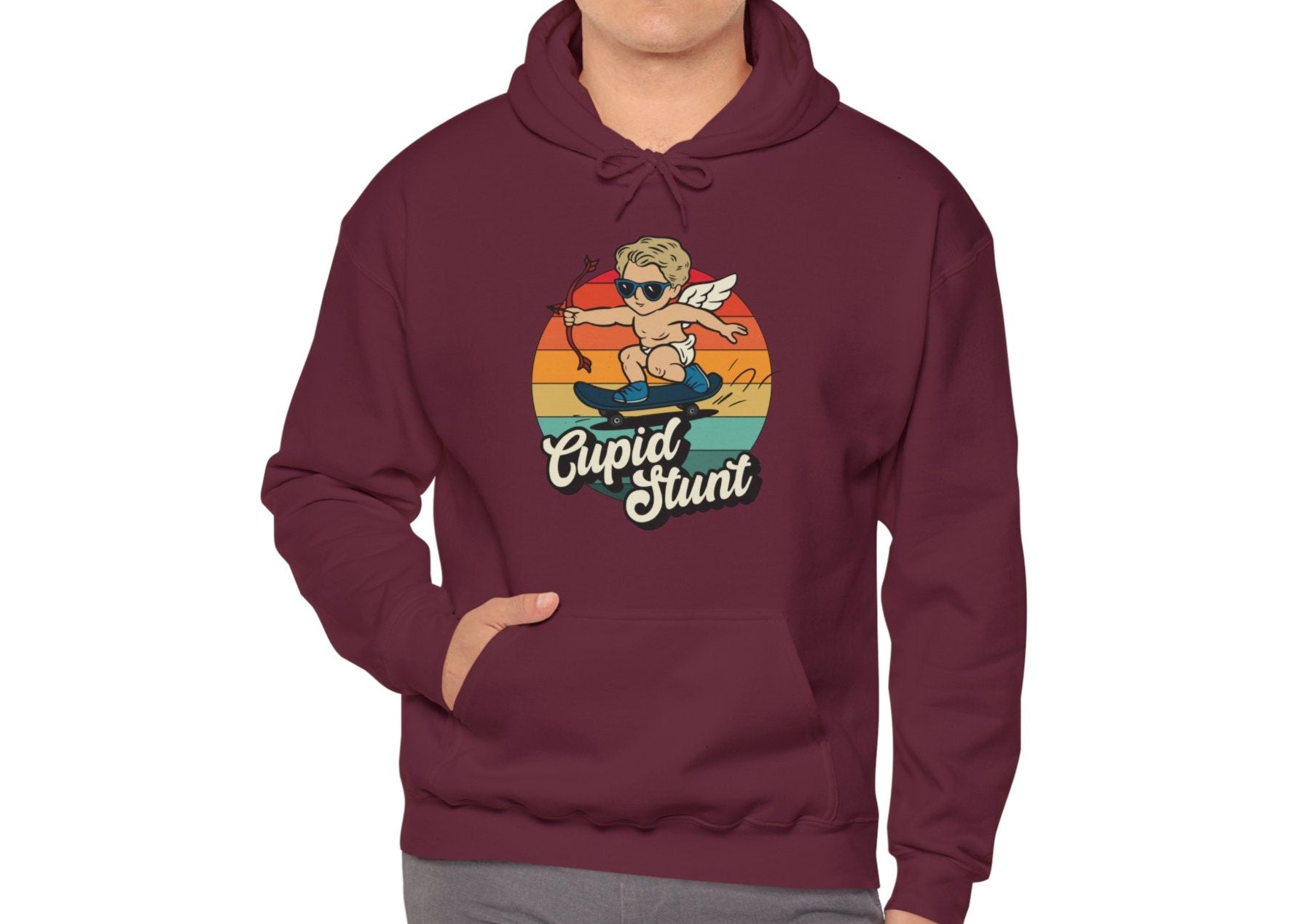Model wearing a maroon hoodie with a retro-style sunset background, an illustration of Cupid riding a skateboard, and the text "Cupid Stunt," standing with hands in pockets. Part of the Vivid Divergence Sensory Friendly Unisex Hoodie Range.