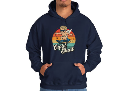 Model wearing a navy hoodie with a retro-style sunset background, an illustration of Cupid riding a skateboard, and the text "Cupid Stunt," standing with hands in pockets. Part of the Vivid Divergence Sensory Friendly Unisex Hoodie Range.