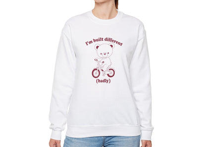 Model wearing a white sweatshirt with a graphic of a bear riding a bike and the text "I'm Built Different (Badly)," standing with hands at sides. Part of the Vivid Divergence Sensory Friendly Unisex Sweater Jumper Range.