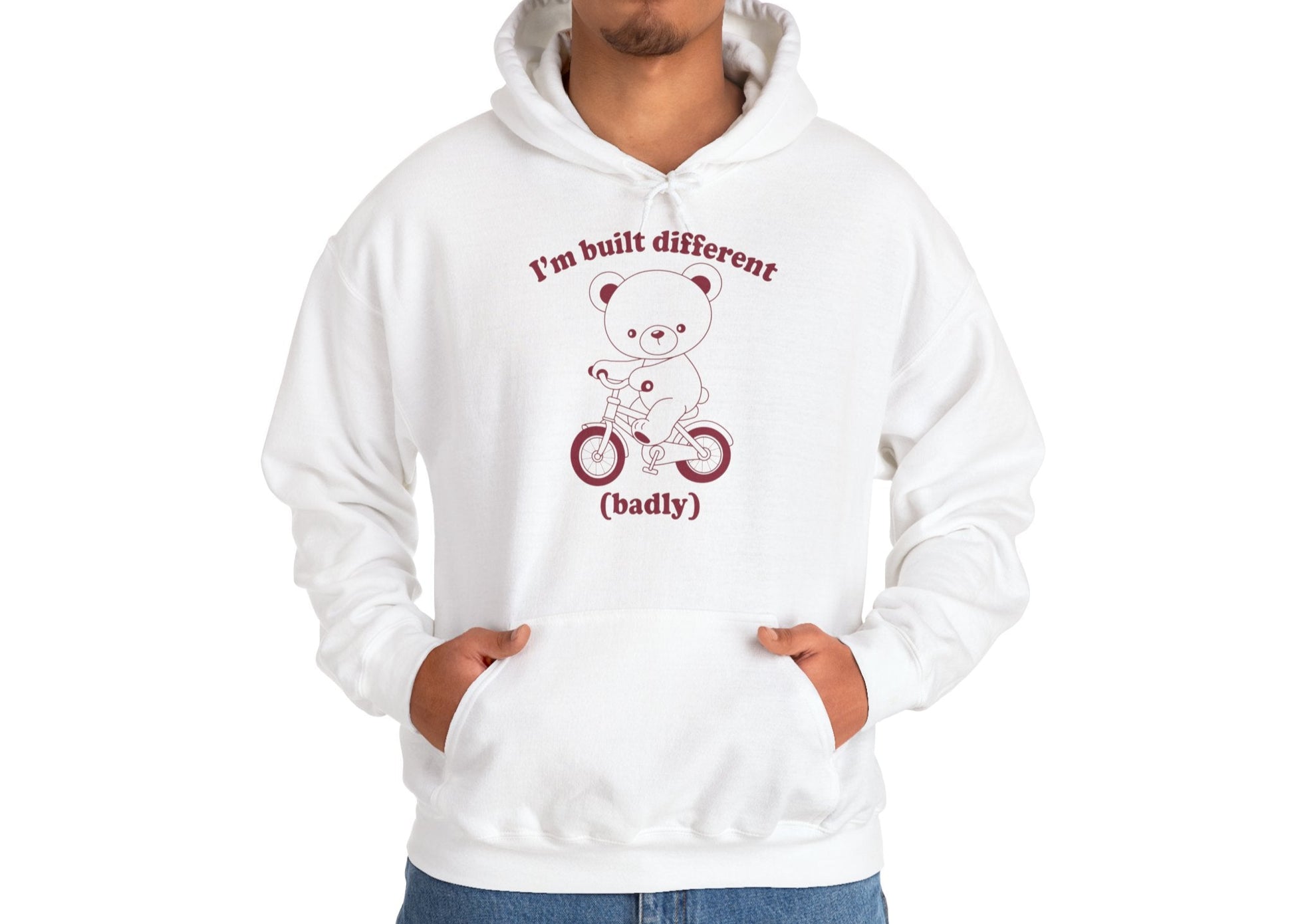 Model wearing a white hoodie with a cartoon bear riding a small bicycle and the text "I'm Built Different (Badly)," standing with hands in pockets. Part of the Vivid Divergence Sensory Friendly Unisex Hoodie Range.