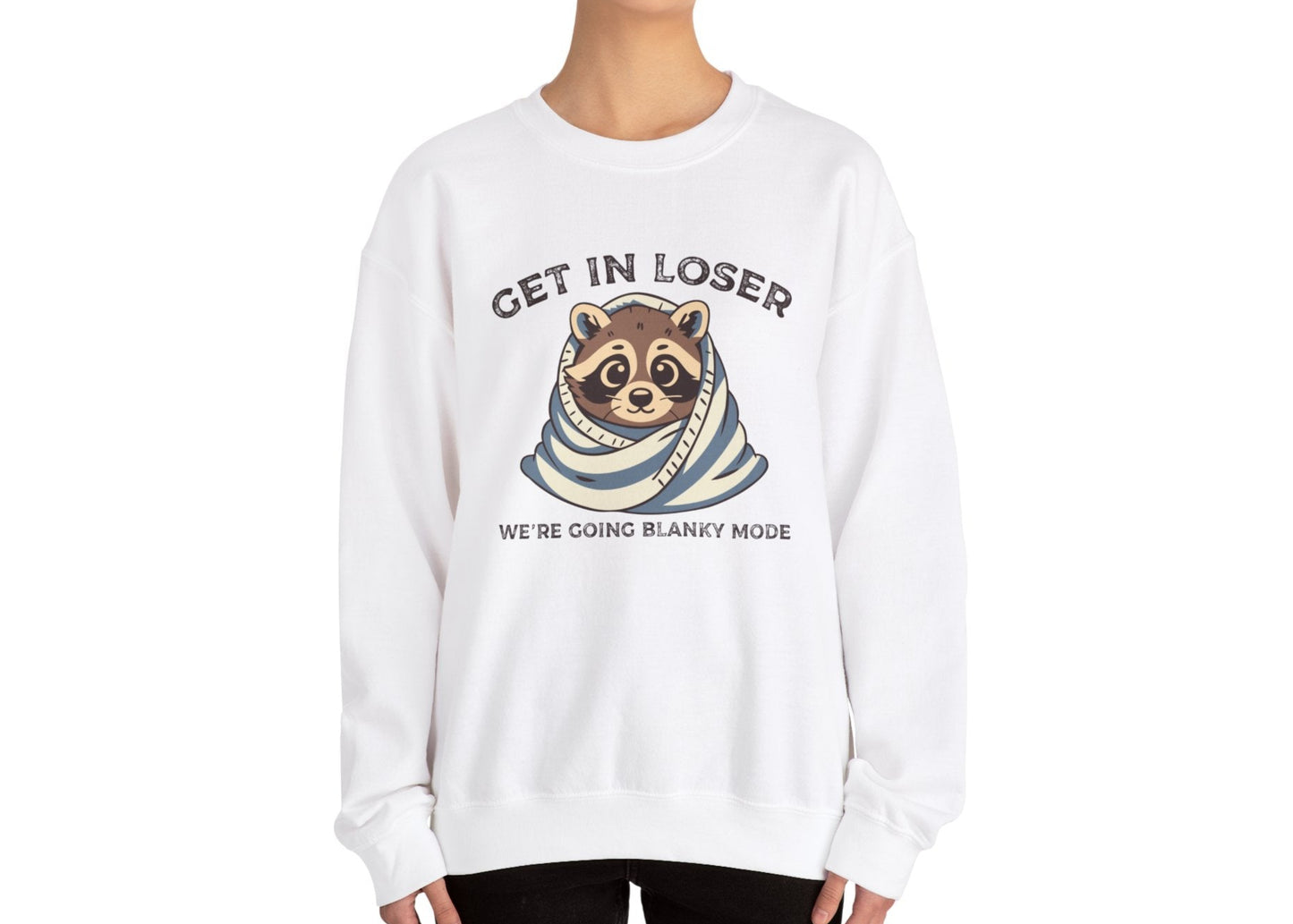 Model wearing a white sweatshirt with a raccoon wrapped in a blanket and the text "Get in Loser, We're Going Blanky Mode," standing with hands at sides. Part of the Vivid Divergence Sensory Friendly Unisex Sweater Jumper Range.