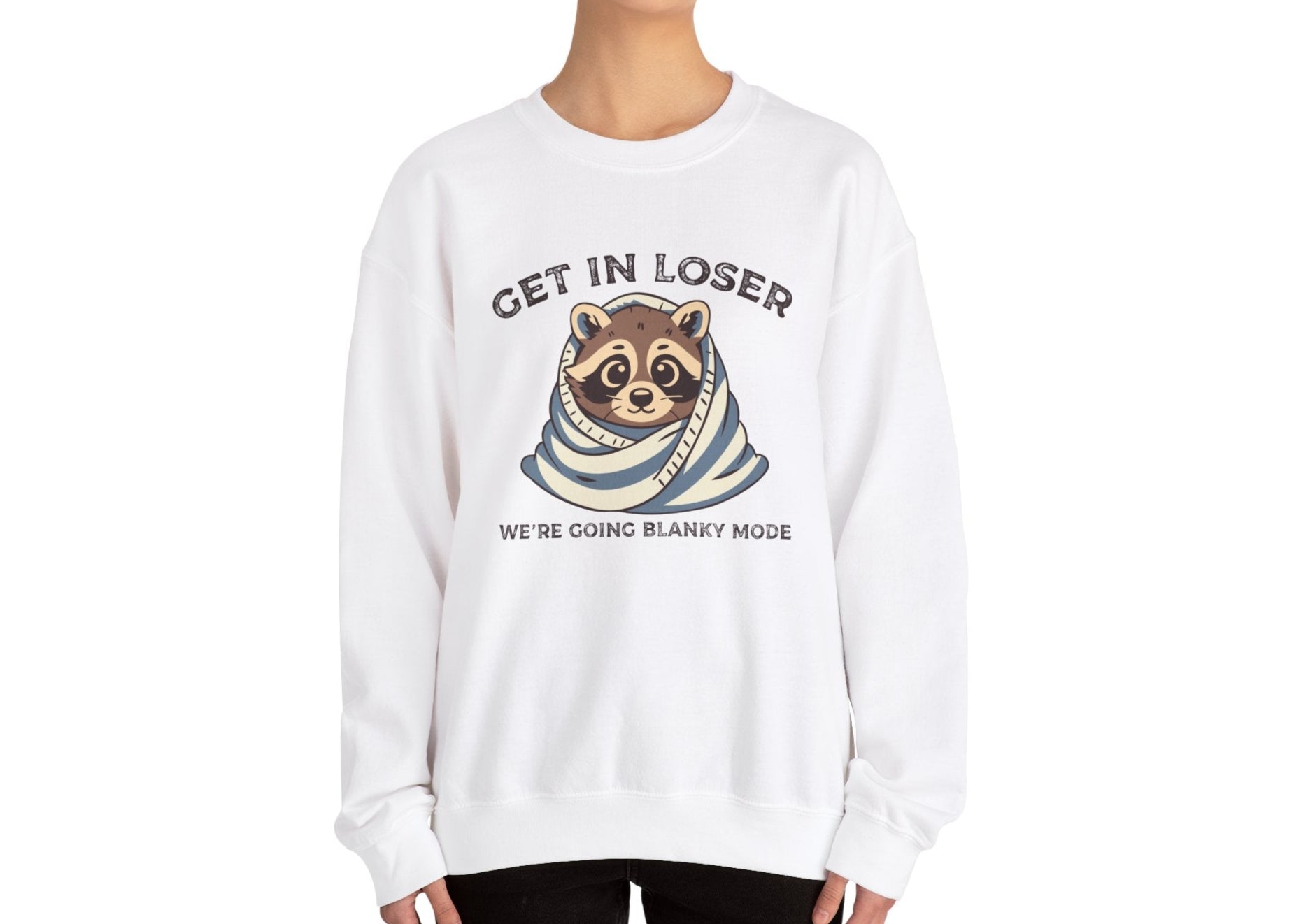 Model wearing a white sweatshirt with a raccoon wrapped in a blanket and the text "Get in Loser, We're Going Blanky Mode," standing with hands at sides. Part of the Vivid Divergence Sensory Friendly Unisex Sweater Jumper Range.