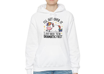 Model wearing a white hoodie with a cartoon unicorn lying dramatically on a couch and the text "I'll Get Over It, I Just Need To Be Dramatic First," standing with hands at sides. Part of the Vivid Divergence Sensory Friendly Unisex Hoodie Range.