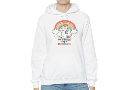 Model wearing a white hoodie with a grumpy-looking unicorn under a rainbow and the text "I'm a Freakin' Ray of Sunshine," standing with hands in pockets. Part of the Vivid Divergence Sensory Friendly Unisex Hoodie Range.