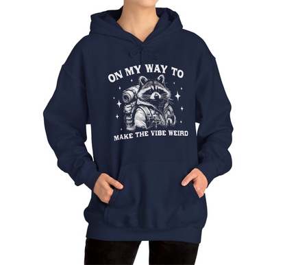 Model wearing a navy blue hoodie featuring the text "On My Way to Make the Vibe Weird" and a cartoon illustration of a raccoon holding a stick. Part of the Vivid Divergence Sensory Friendly Unisex Hoodie Range.