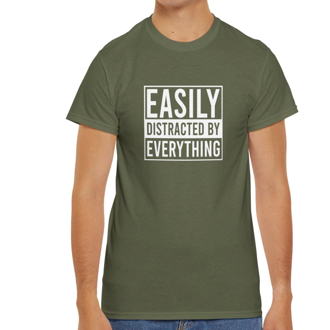 A person wearing an olive green T-shirt with "Easily Distracted by Everything" text, standing casually. Part of the Vivid Divergence Sensory Friendly Unisex Tees Range.