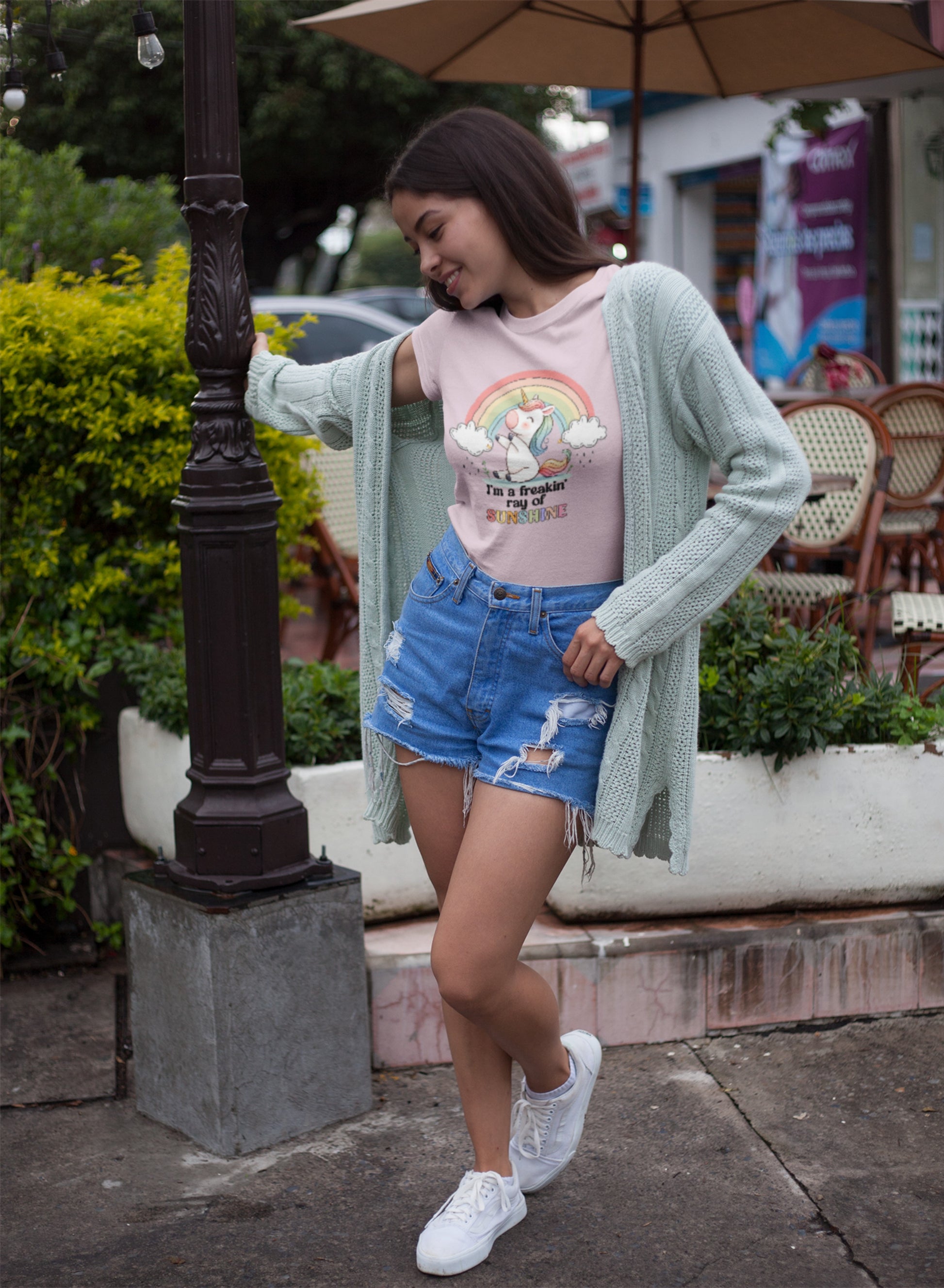 A person outdoors wearing a white T-shirt with a rainbow and unicorn graphic and the text "I'm A Freakin Ray Of Sunshine." Part of the Vivid Divergence Sensory Friendly Unisex Tees Range.