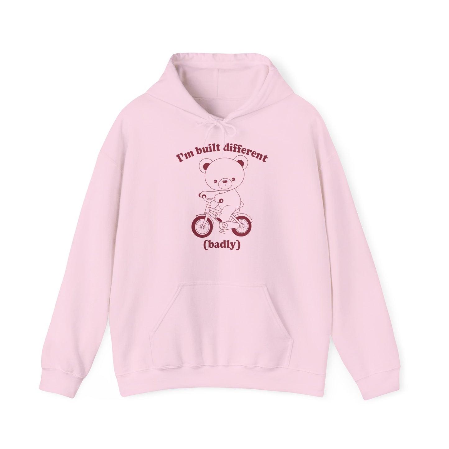 Front view of a pink hoodie featuring a cartoon bear riding a small bicycle with the text "I'm Built Different (Badly)." Part of the Vivid Divergence Sensory Friendly Unisex Hoodie Range.