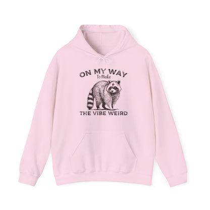 Front view of a pink hoodie featuring the text "On My Way to Make the Vibe Weird" and a cartoon illustration of a raccoon holding a stick. Part of the Vivid Divergence Sensory Friendly Unisex Hoodie Range.