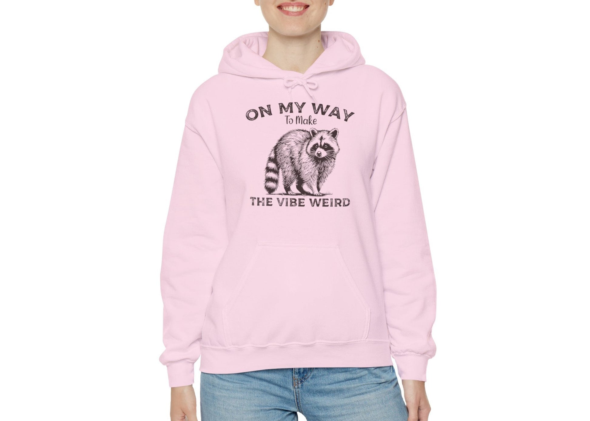 Model wearing a pink hoodie featuring the text "On My Way to Make the Vibe Weird" and a cartoon illustration of a raccoon holding a stick. Part of the Vivid Divergence Sensory Friendly Unisex Hoodie Range.