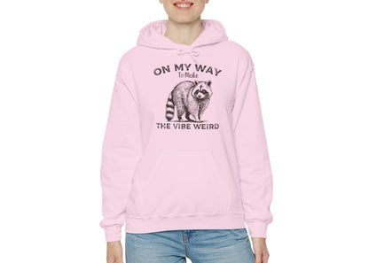 Model wearing a pink hoodie featuring the text "On My Way to Make the Vibe Weird" and a cartoon illustration of a raccoon holding a stick. Part of the Vivid Divergence Sensory Friendly Unisex Hoodie Range.