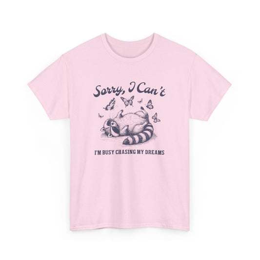 Front view of a pink T-shirt with "Sorry I Can't, I'm Busy Chasing My Dreams" and a playful sleeping raccoon graphic in black text. Part of the Vivid Divergence Sensory Friendly Unisex Tees Range.