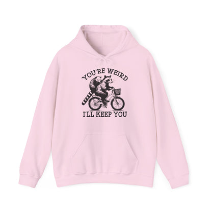 Front view of a pink hoodie featuring the text "You're Weird, I'll Keep You" with a cartoon illustration of two raccoons riding a bicycle. Part of the Vivid Divergence Sensory Friendly Unisex Hoodie Range.