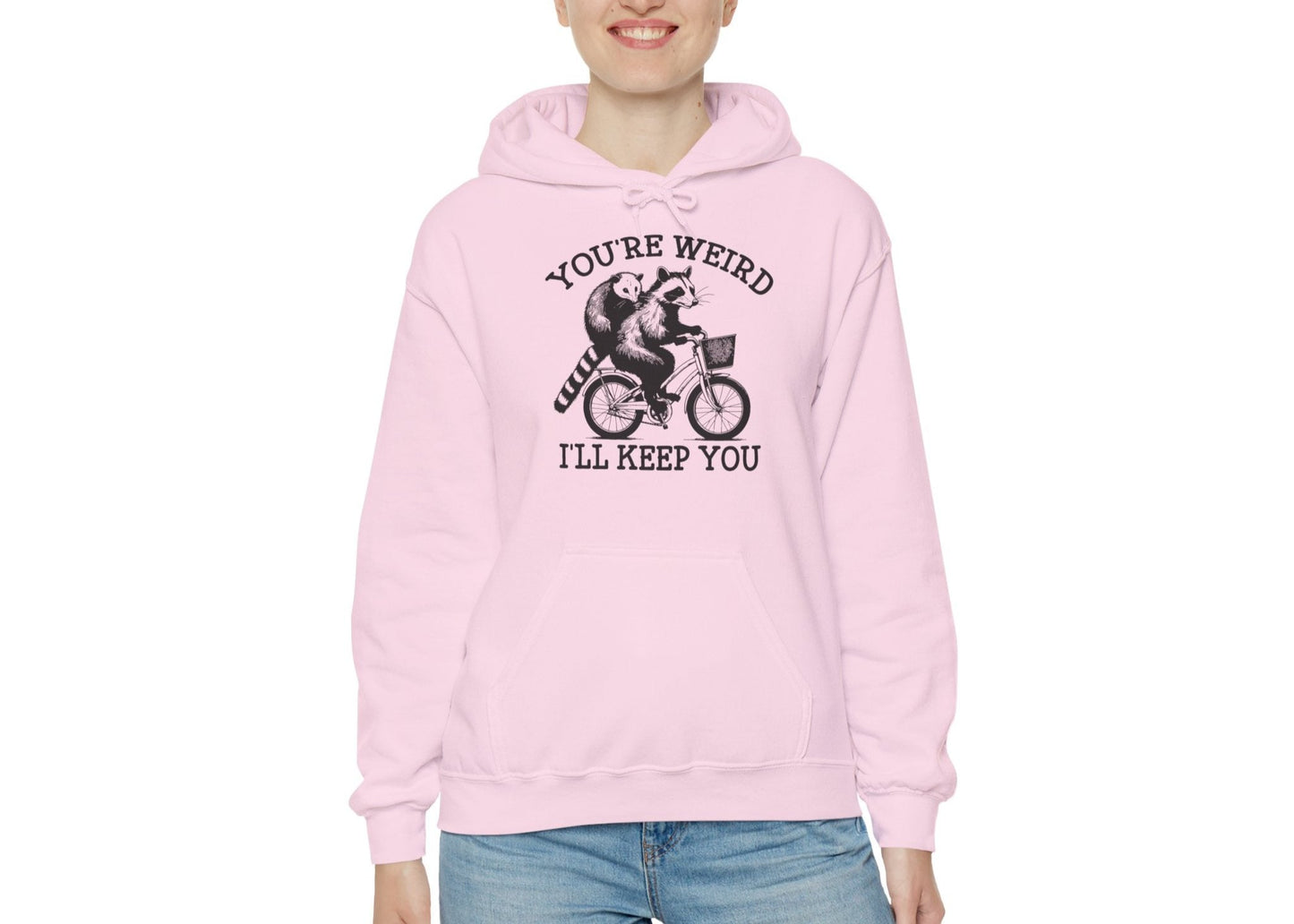 Model wearing a pink hoodie featuring the text "You're Weird, I'll Keep You" with a cartoon illustration of two raccoons riding a bicycle. Part of the Vivid Divergence Sensory Friendly Unisex Hoodie Range.