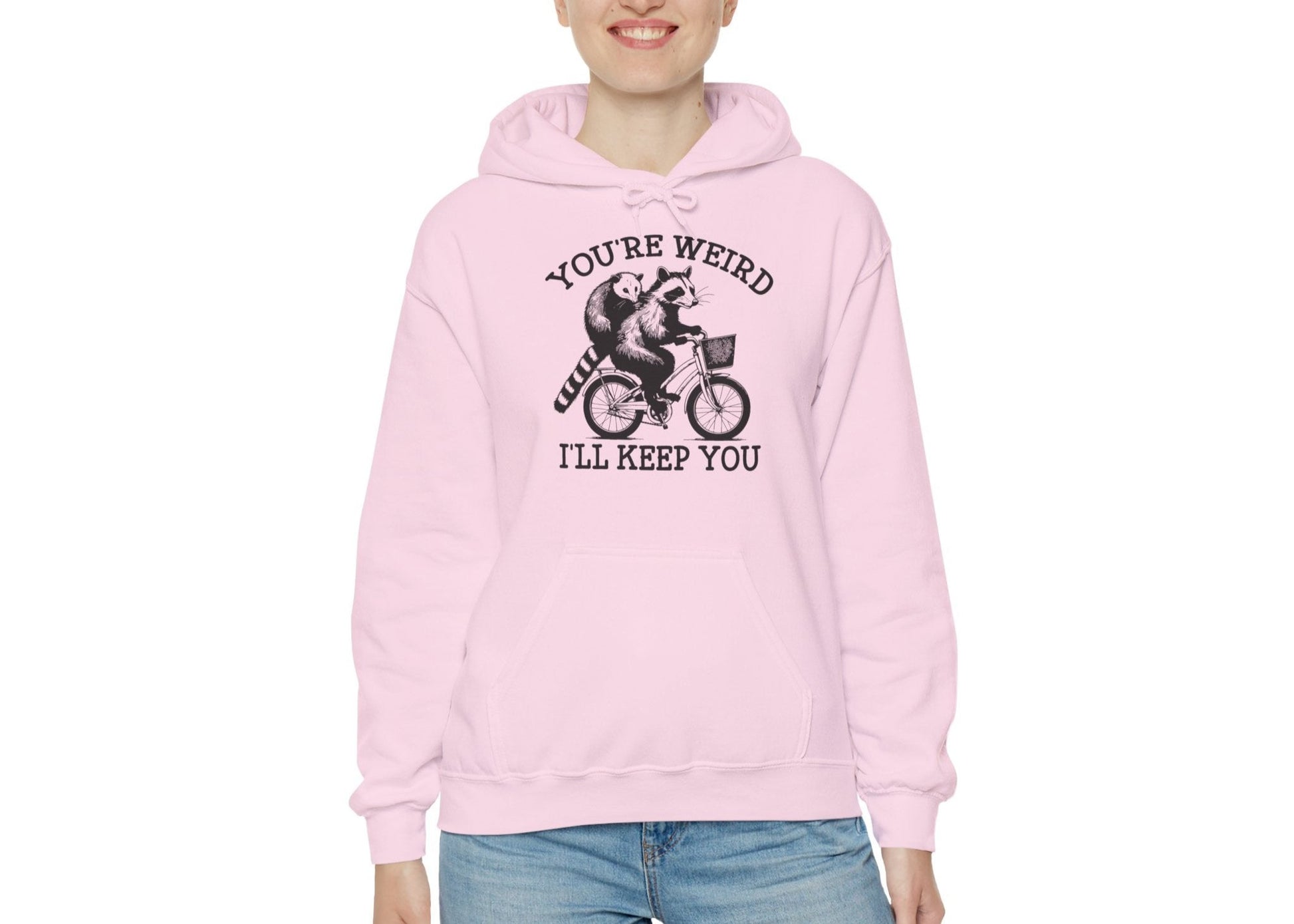 Model wearing a pink hoodie featuring the text "You're Weird, I'll Keep You" with a cartoon illustration of two raccoons riding a bicycle. Part of the Vivid Divergence Sensory Friendly Unisex Hoodie Range.