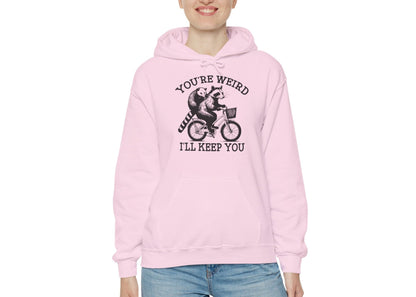 Model wearing a pink hoodie featuring the text "You're Weird, I'll Keep You" with a cartoon illustration of two raccoons riding a bicycle. Part of the Vivid Divergence Sensory Friendly Unisex Hoodie Range.