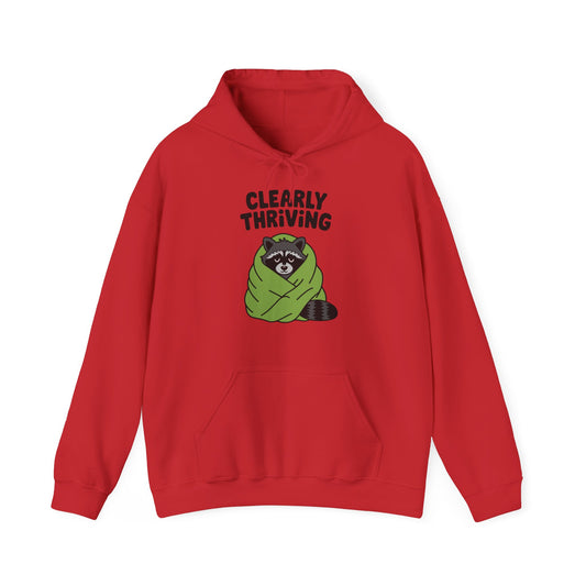 Front view of a red hoodie featuring the text "Clearly Thriving" and a cartoon illustration of a raccoon wrapped in a cozy blanket. Part of the Vivid Divergence Sensory Friendly Unisex Hoodie Range.