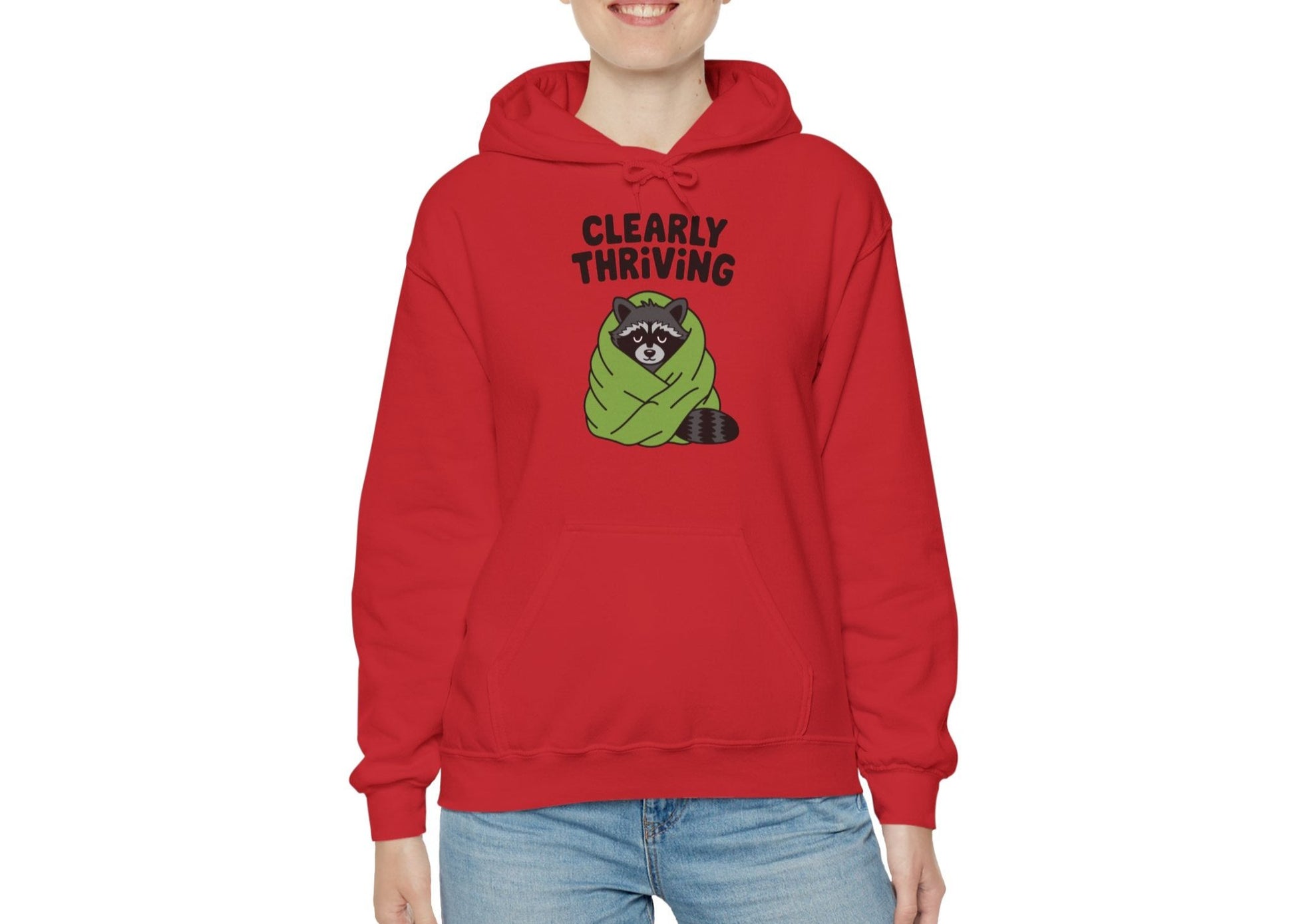 Model wearing a red hoodie featuring the text "Clearly Thriving" and a cartoon illustration of a raccoon wrapped in a cozy blanket. Part of the Vivid Divergence Sensory Friendly Unisex Hoodie Range.