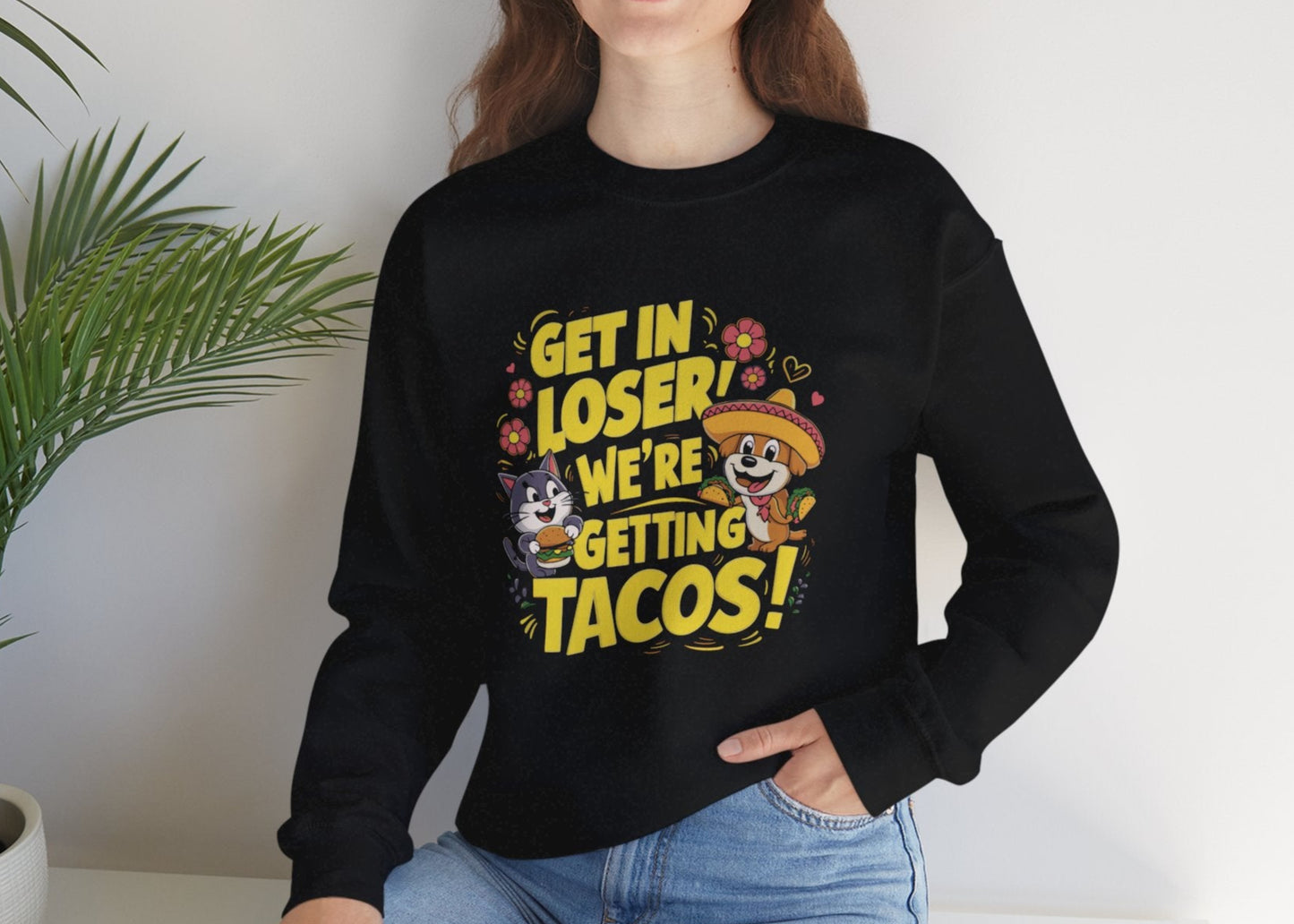 Model seated, wearing a black sweatshirt with colorful cartoon animals and the text "Get in Loser, We're Getting Tacos!" Part of the Vivid Divergence Sensory Friendly Unisex Sweater Jumper Range.