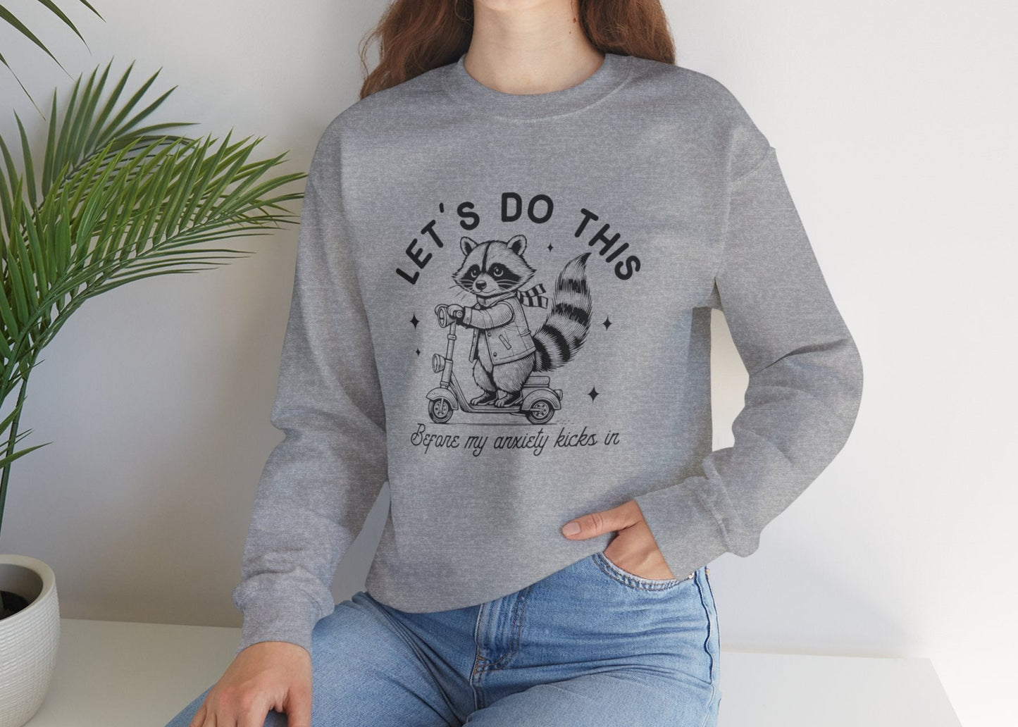 Model seated, wearing a gray sweatshirt with a cartoon raccoon riding a scooter and the text "Let's Do This Before My Anxiety Kicks In." Part of the Vivid Divergence Sensory Friendly Unisex Sweater Jumper Range.
