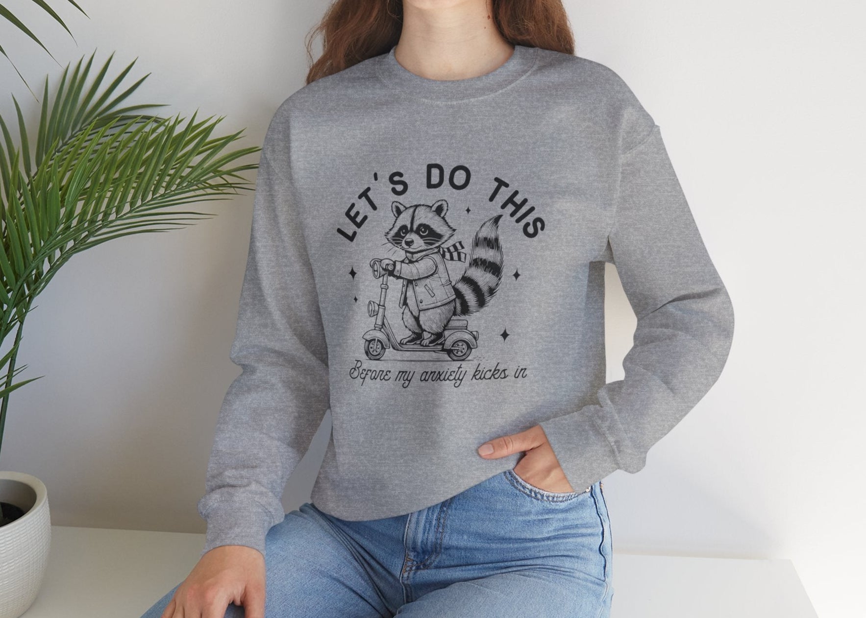 Model seated, wearing a gray sweatshirt with a cartoon raccoon riding a scooter and the text "Let's Do This Before My Anxiety Kicks In." Part of the Vivid Divergence Sensory Friendly Unisex Sweater Jumper Range.