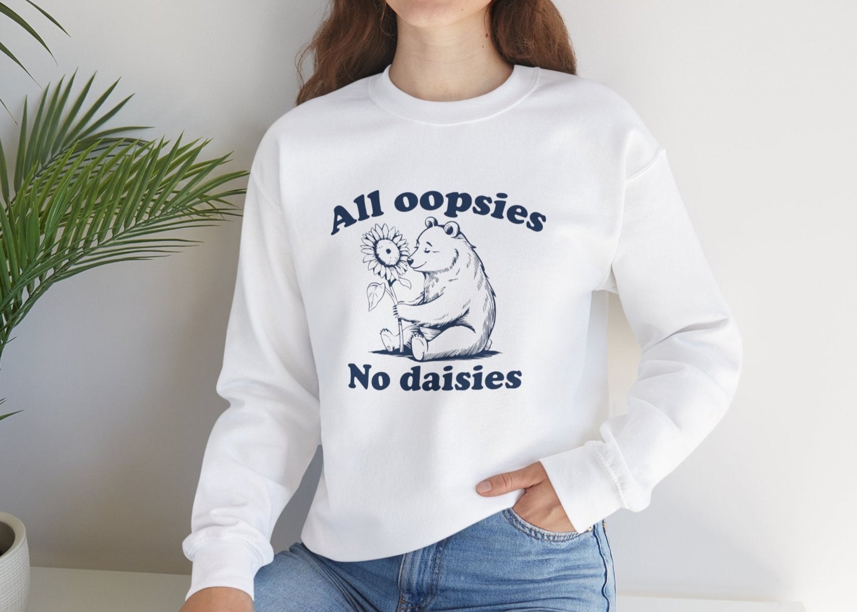 Model seated, wearing a white sweatshirt with a cartoon bear holding a sunflower and the text "All Oopsies, No Daisies." Part of the Vivid Divergence Sensory Friendly Unisex Sweater Jumper Range.